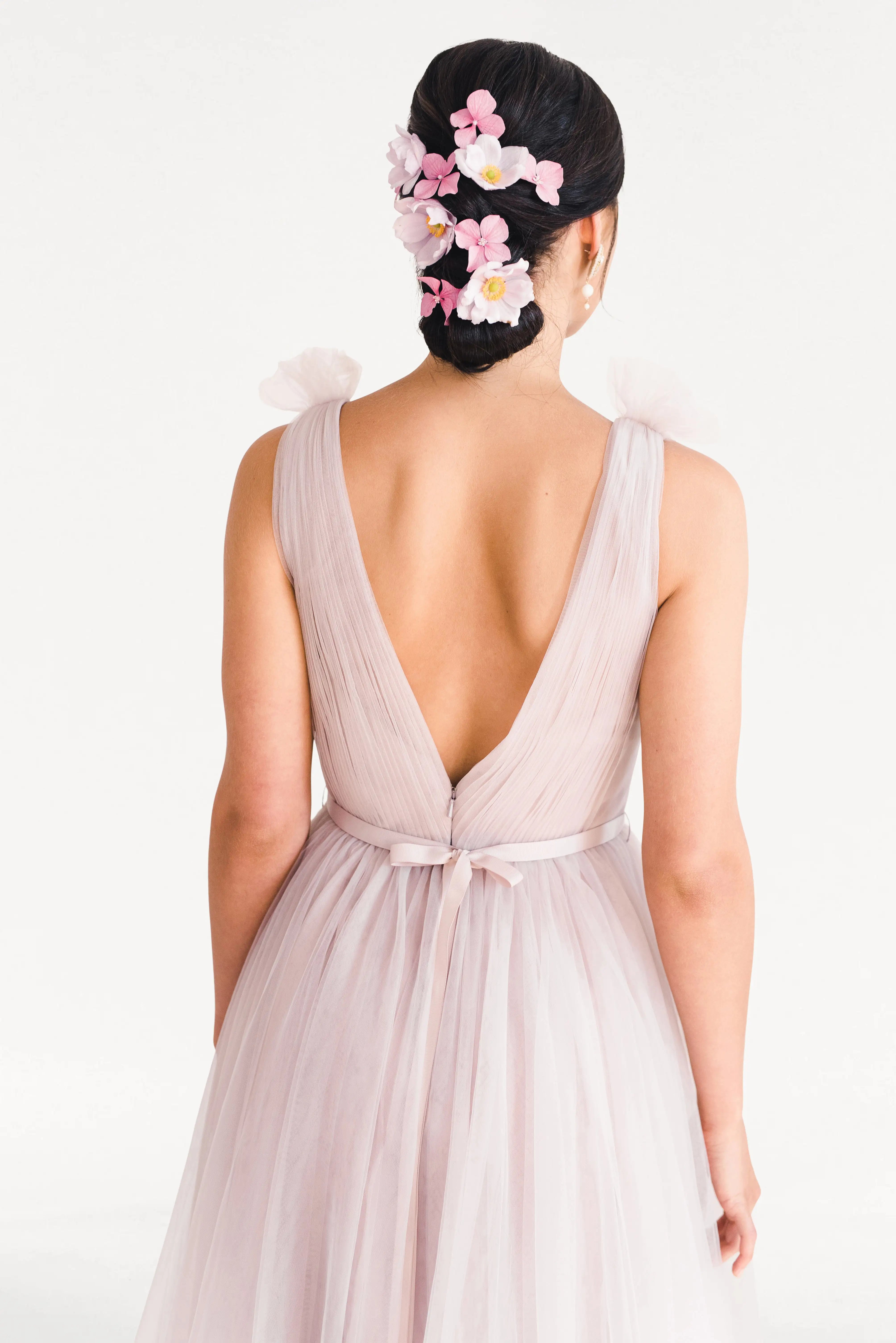 A woman faces away in the TH&TH Grace Bridesmaid Dress in Smoked Blush Tulle, a light pink sleeveless gown with a deep V back. Her dark hair is in a low bun adorned with pink and white flower clips, against a plain white background.
