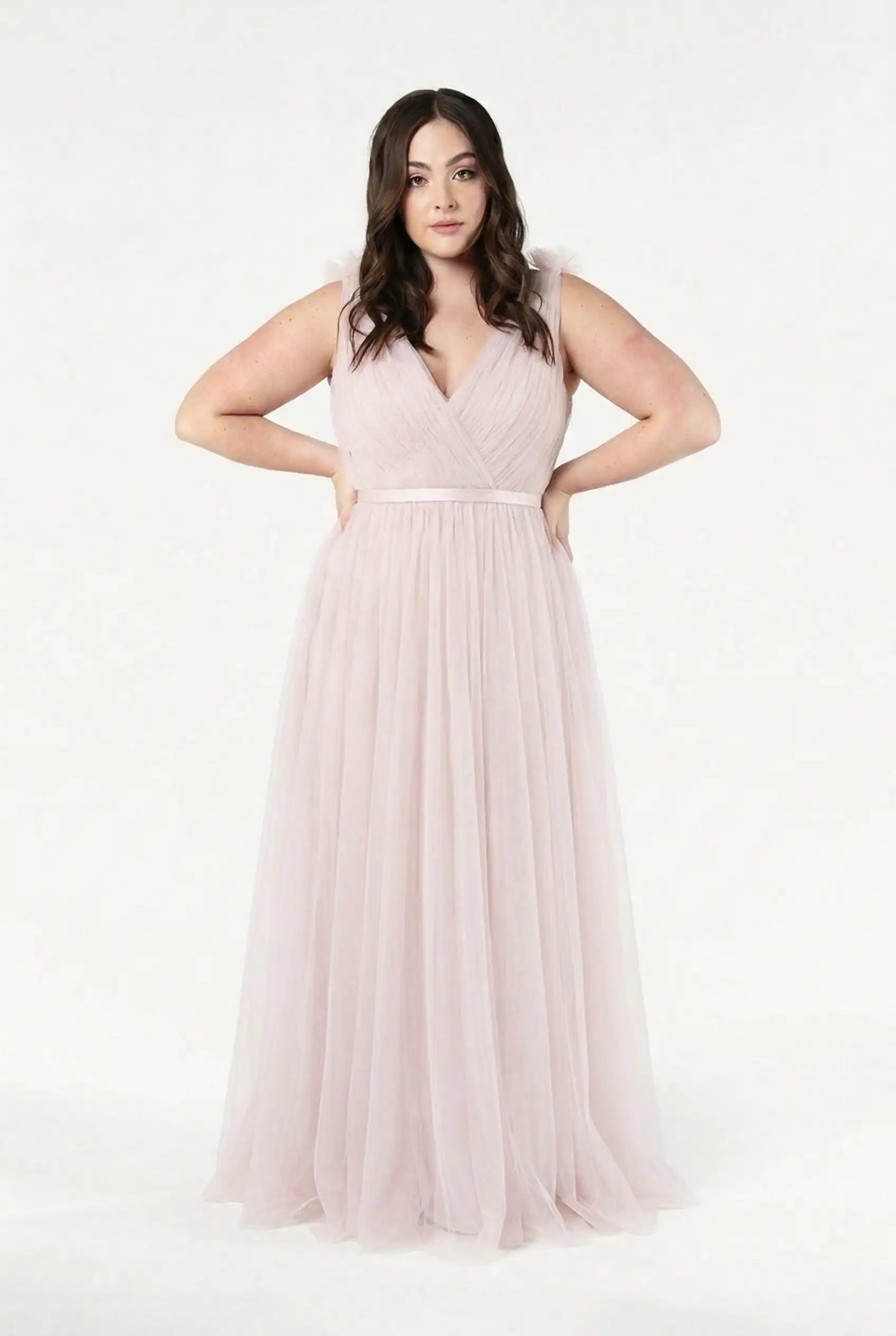 A woman with long, wavy brown hair models the TH&TH Grace Bridesmaid Dress in Smoked Blush Tulle, a light pink, floor-length gown with a V-neckline—an ideal pick for a romantic bridal party.