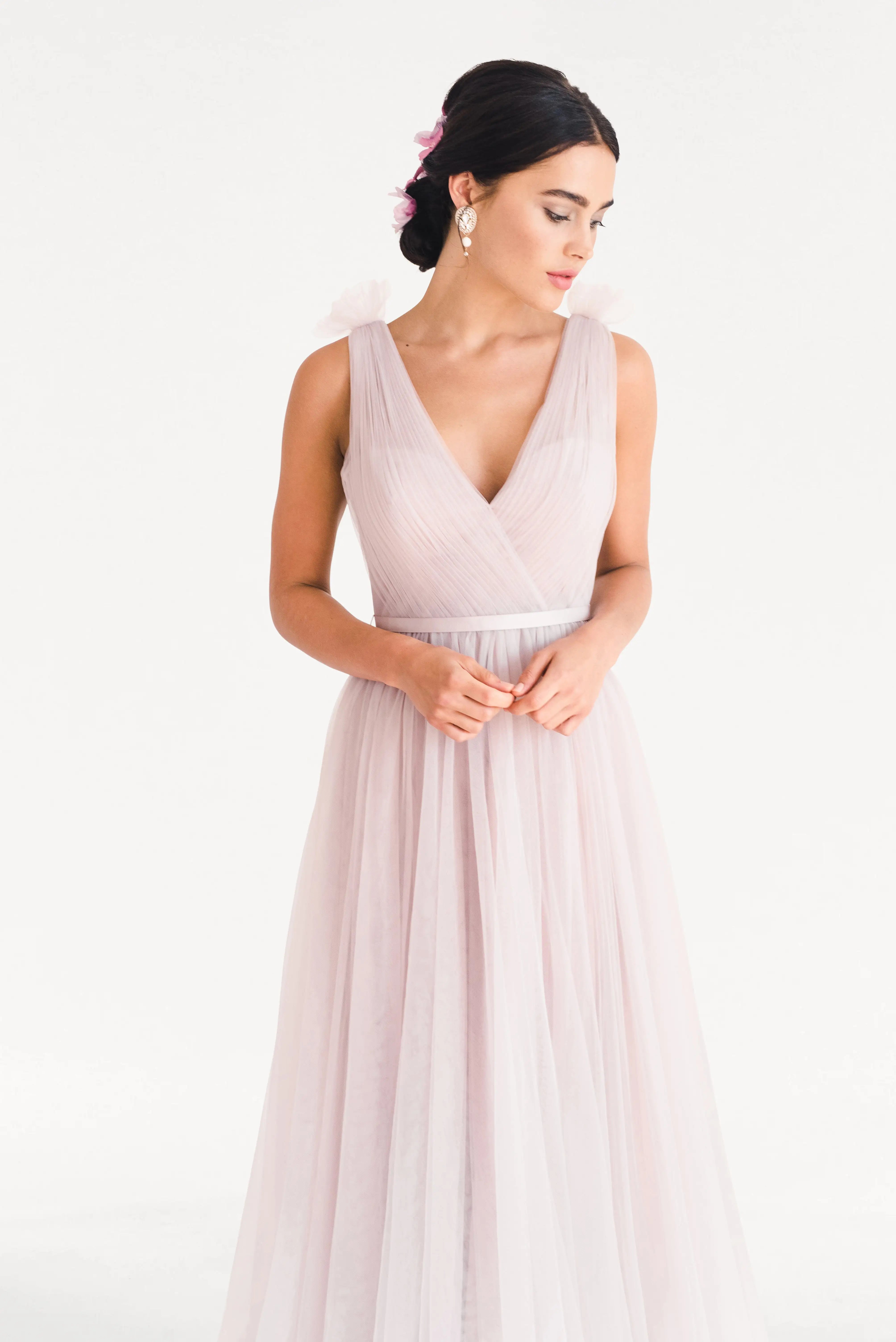 A woman stands against a white background wearing the TH&TH Grace Bridesmaid Dress in Smoked Blush Tulle, a sleeveless gown with a V-neckline. Pink flowers and drop earrings complete this romantic look, perfect for any bridal party.