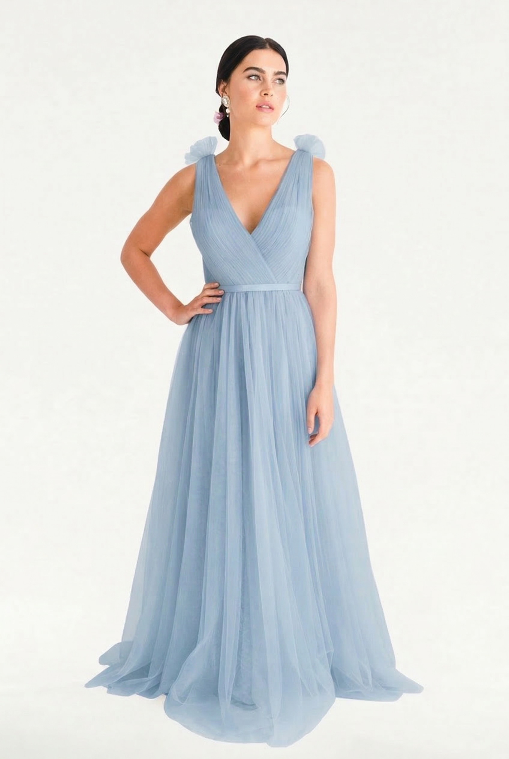 A woman models the TH&TH Grace Bridesmaid Dress in Dusty Blue Tulle, a sleeveless, floor-length gown with a V-neckline and soft shoulder details, standing against a white background with one hand on her hip.
