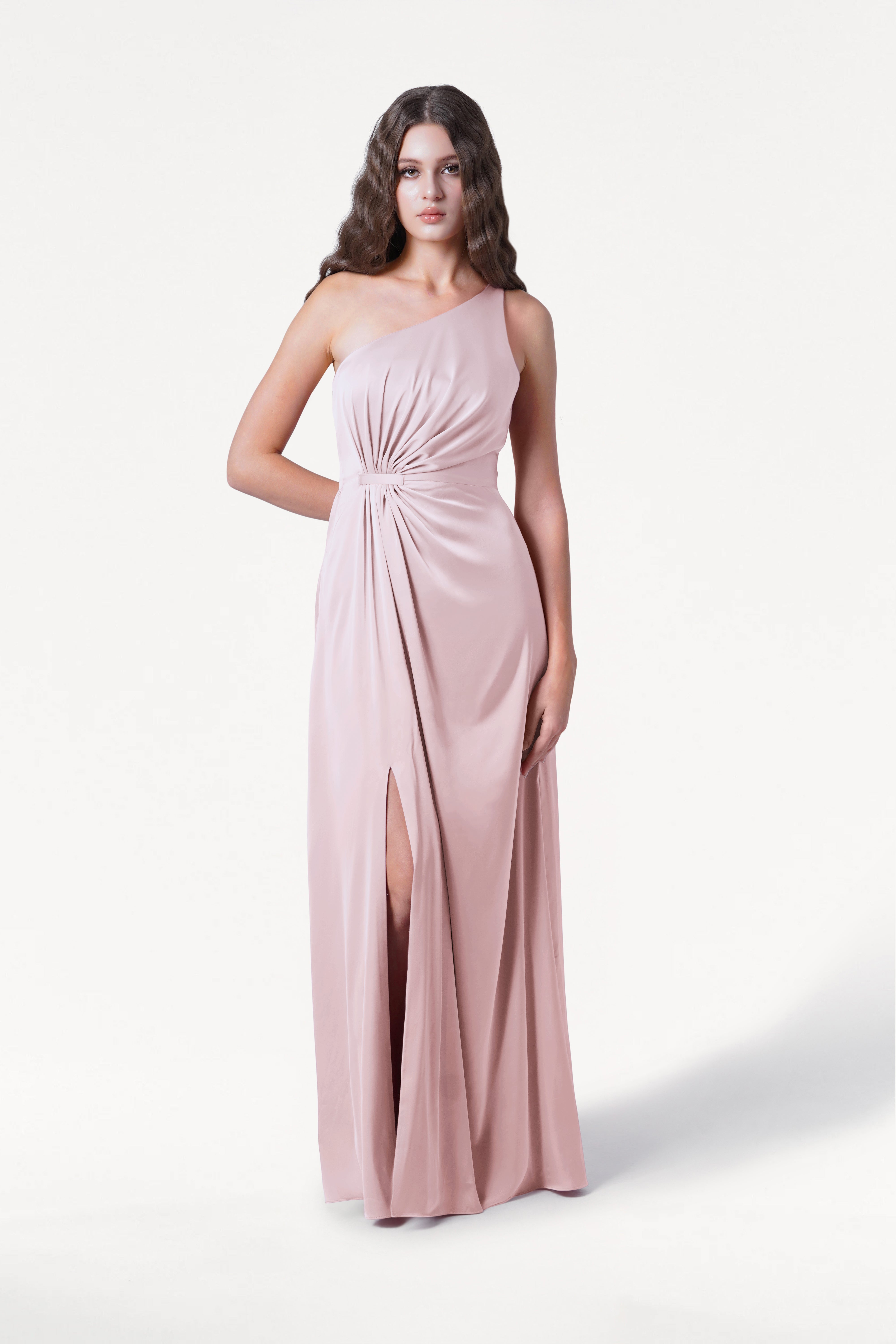 A woman wears the TH&TH Greta Bridesmaid Dress in blush pink satin, featuring a one-shoulder design, Grecian draping, gathered knot at the waist, and front slit, standing against a plain white background.