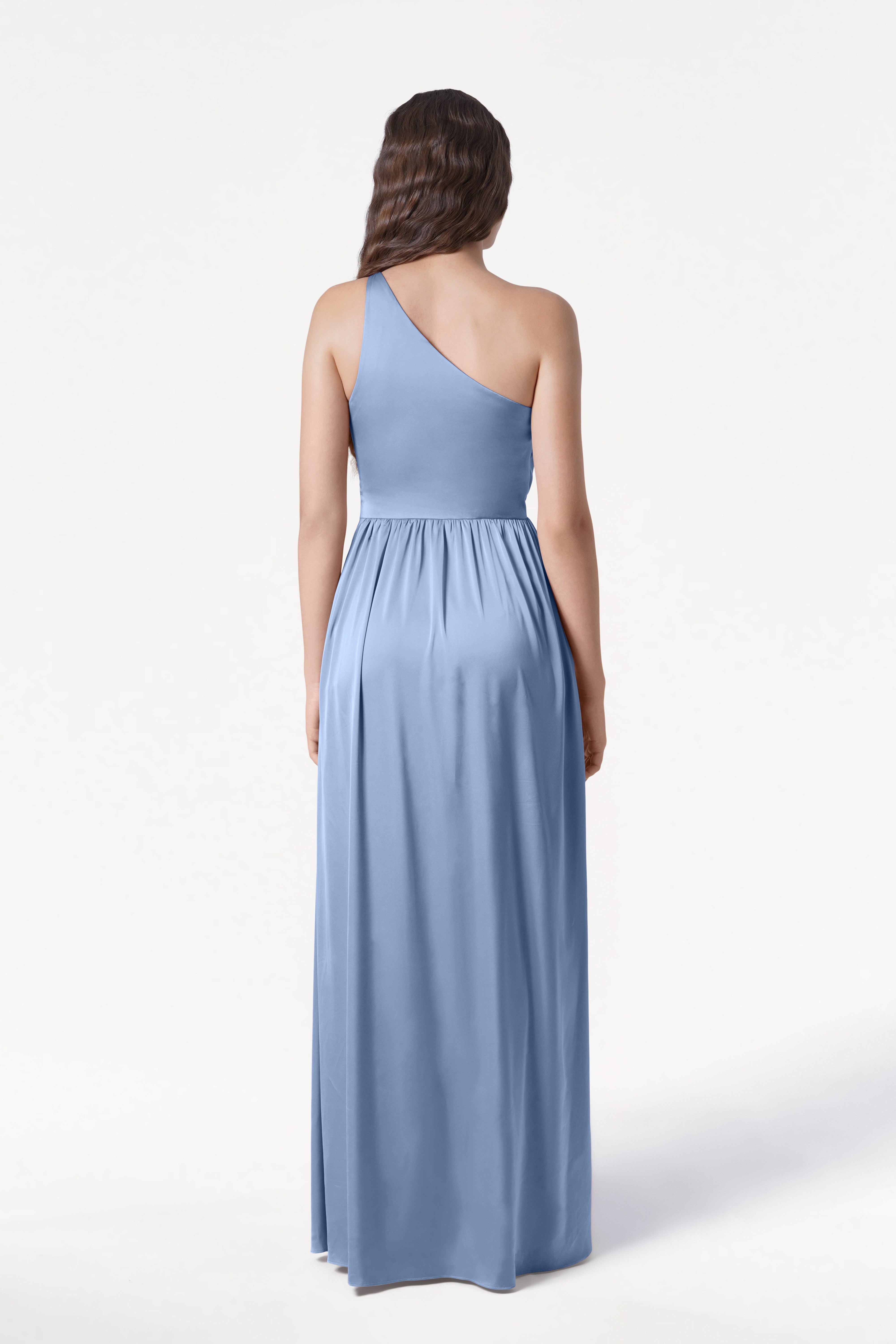 A woman with long, wavy brown hair is seen from the back wearing the TH&TH Greta Bridesmaid Dress in Dusty Blue Satin, a floor-length one-shoulder gown with elegant Grecian draping against a plain white background.