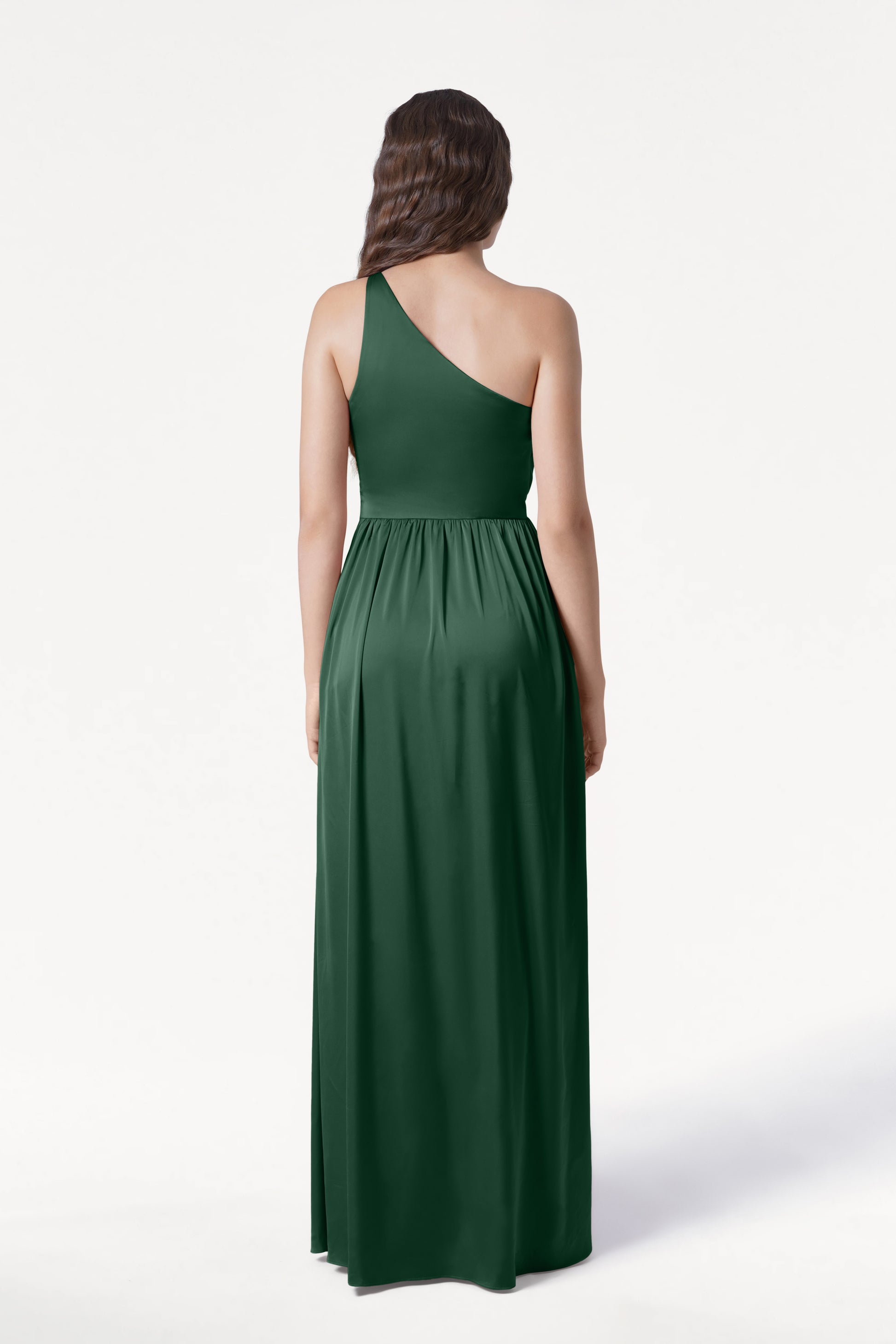 A woman faces away from the camera, showcasing the Greta Bridesmaid Dress in Olive Grove Satin by TH&TH—a floor-length, dark green one-shoulder gown with elegant Grecian draping. Her long, wavy brown hair stands out against a plain white background.
