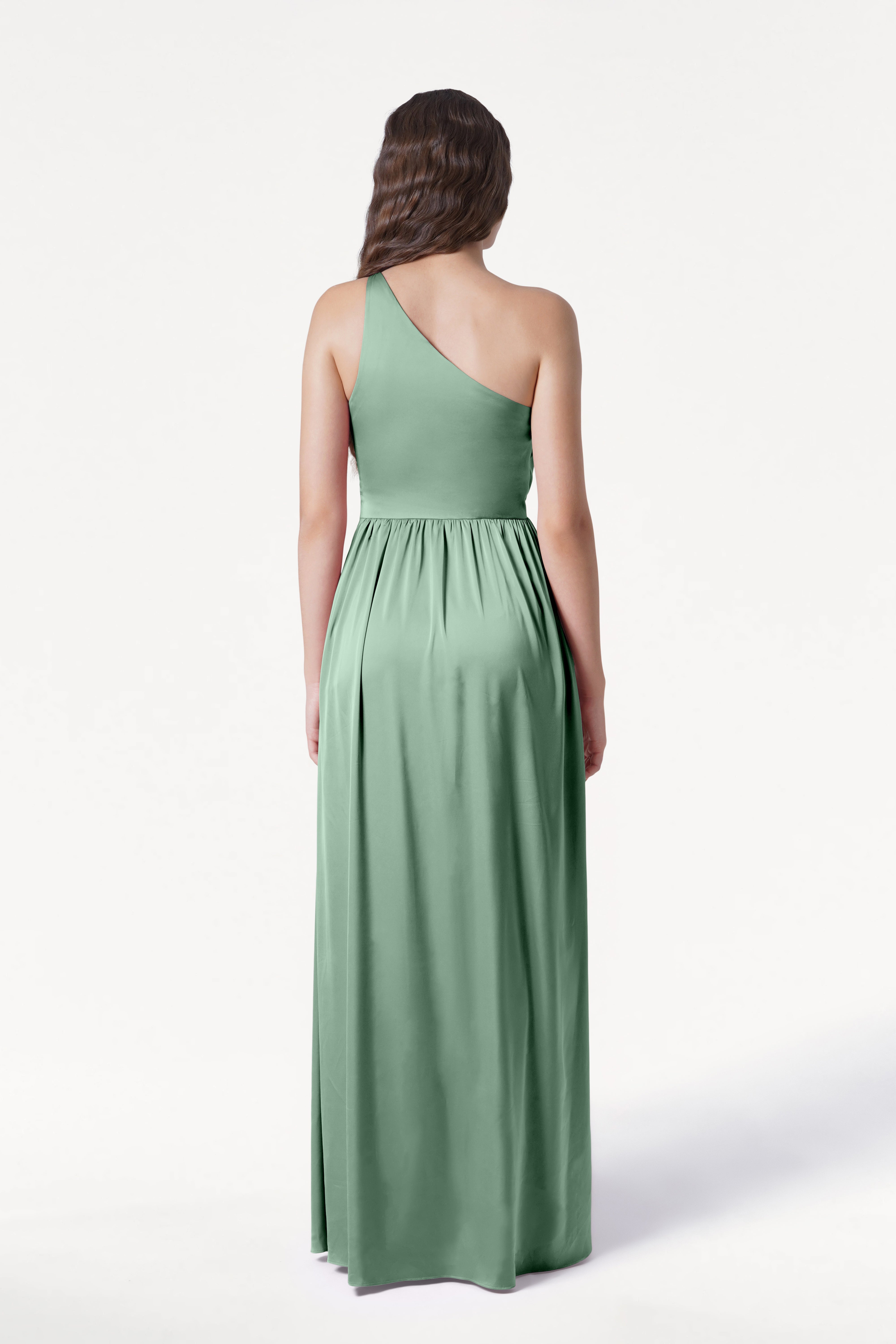 A woman with long, wavy brown hair models the TH&TH Greta Bridesmaid Dress in sage green satin, featuring elegant Grecian draping. She stands with her back to the camera against a plain white background.