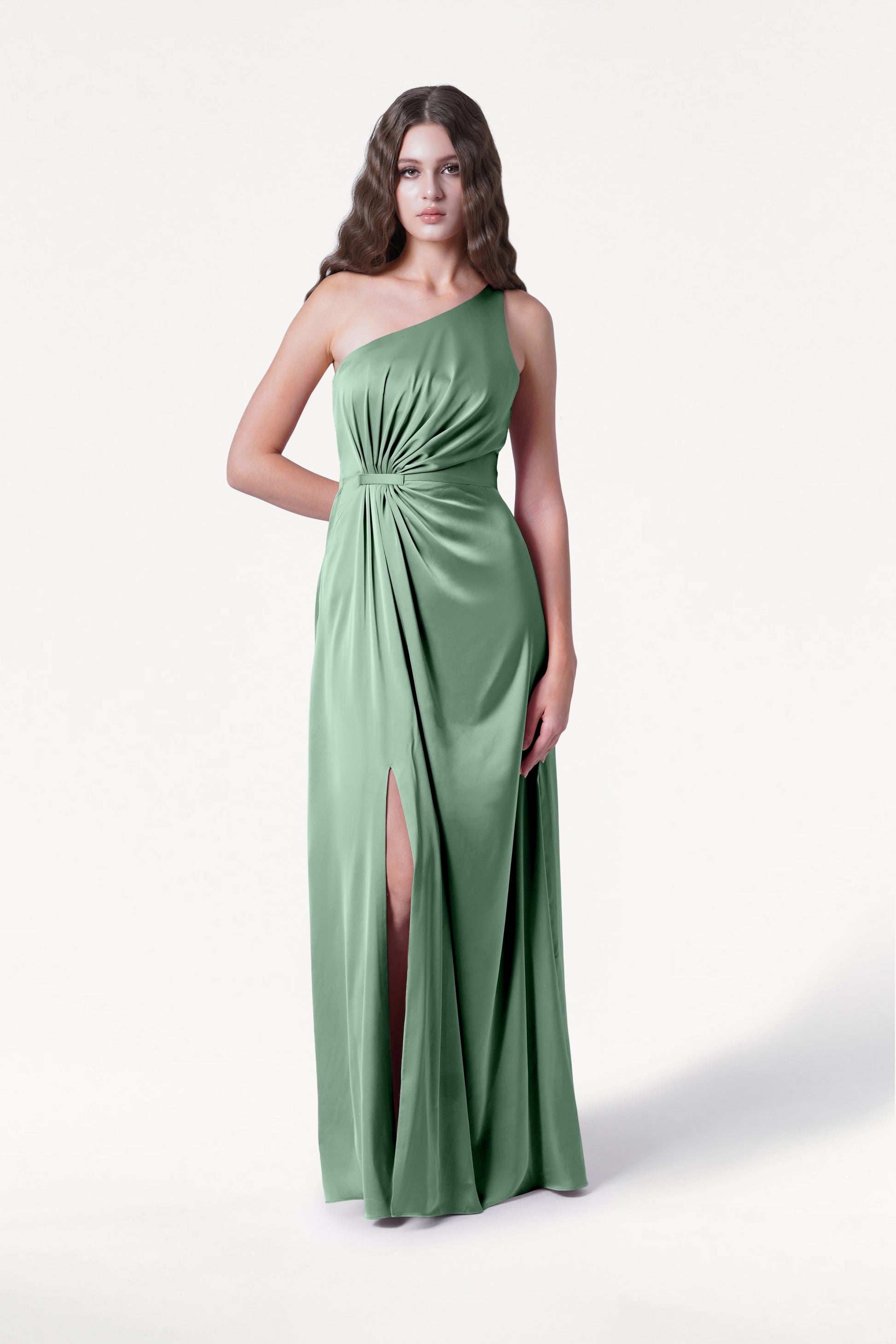 A woman with long wavy hair wears the TH&TH Greta Bridesmaid Dress in Sage Green Satin, a one-shoulder gown with Grecian draping, a gathered waist knot, and a thigh-high slit, standing against a plain white background.