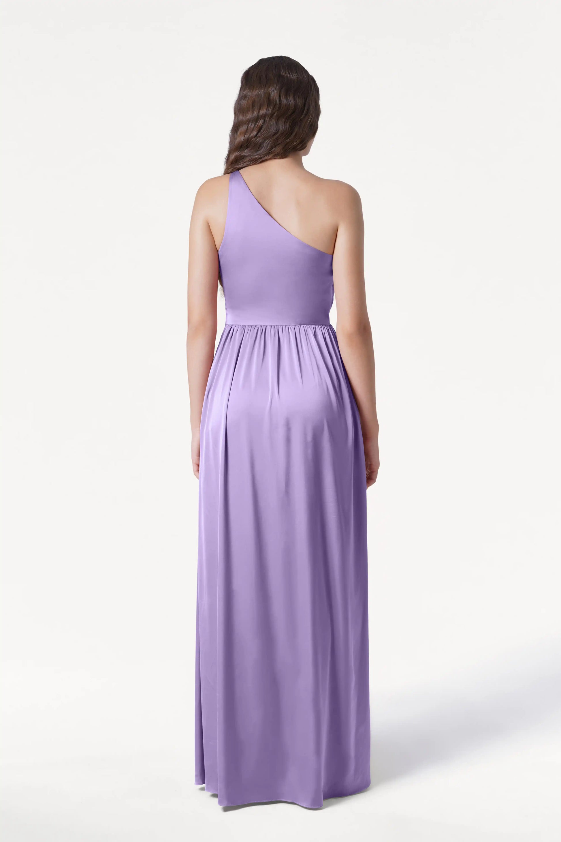 A woman with loose, wavy hair faces away, wearing the TH&TH Greta Bridesmaid Dress in violet satin—a long, sleeveless one-shoulder style with Grecian draping, gathered waist, and flowing skirt—set against a plain white background.