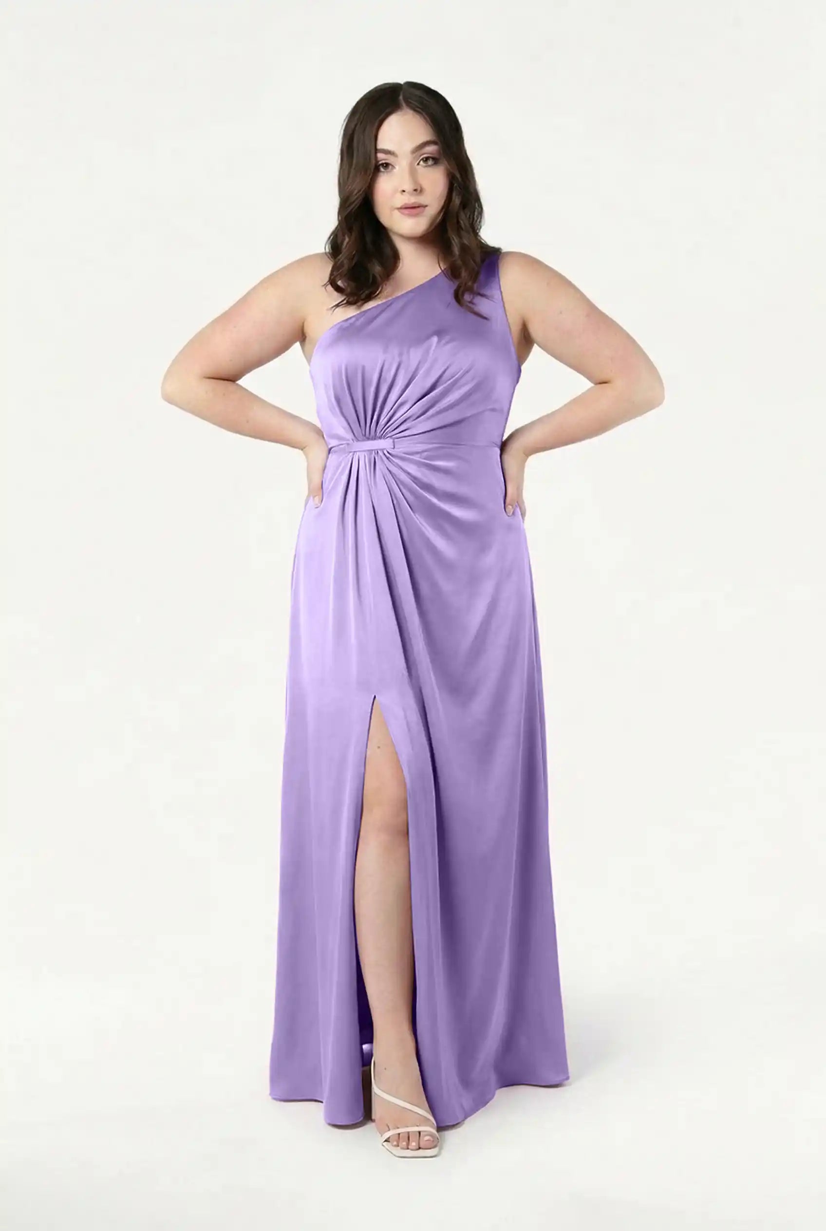 A woman models the TH&TH Greta Bridesmaid Dress in violet satin—a sleeveless, one-shoulder gown with Grecian draping, a gathered waist, and thigh-high slit. She pairs it with white strappy heels against a plain light background.