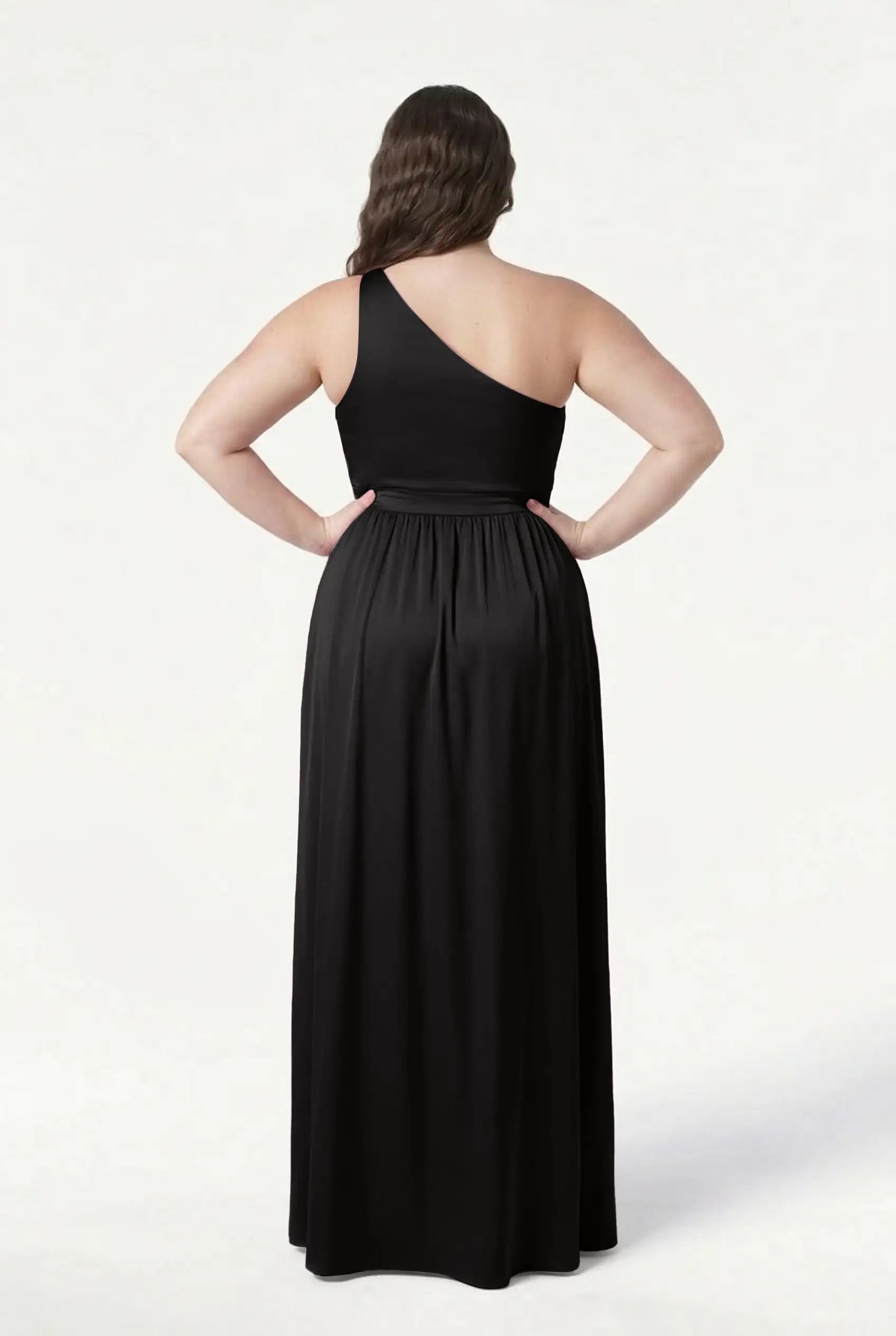 A woman stands with her back to the camera, hands on hips, modeling the TH&TH Greta Bridesmaid Dress in Black Satin, featuring a one-shoulder design, fitted waist, and flowing skirt against a plain white background.