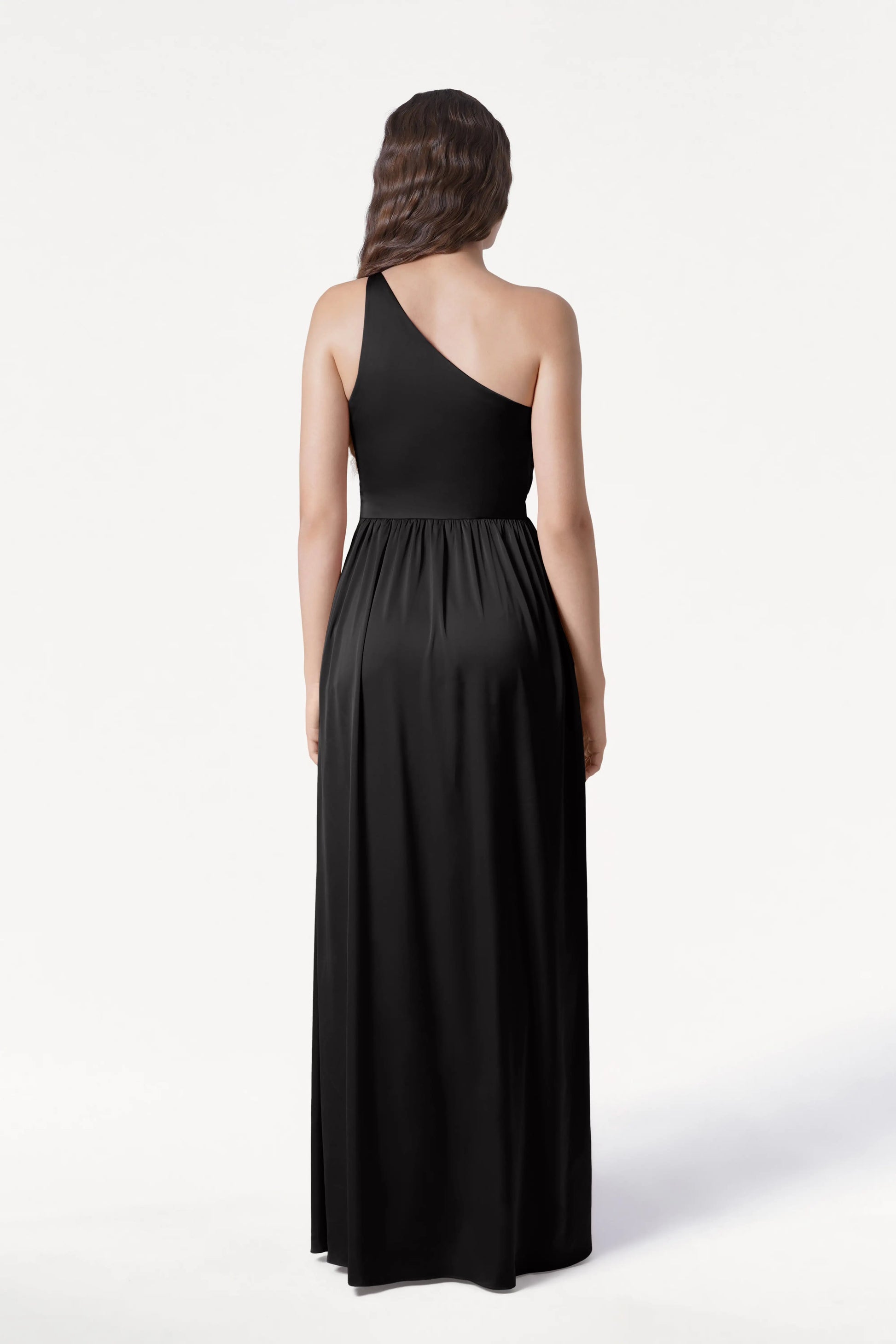A woman with long brown hair, seen from the back, wears the TH&TH Greta Bridesmaid Dress in Black Satin—an elegant, asymmetrical one-shoulder gown with Grecian side draping—posed against a plain white background.