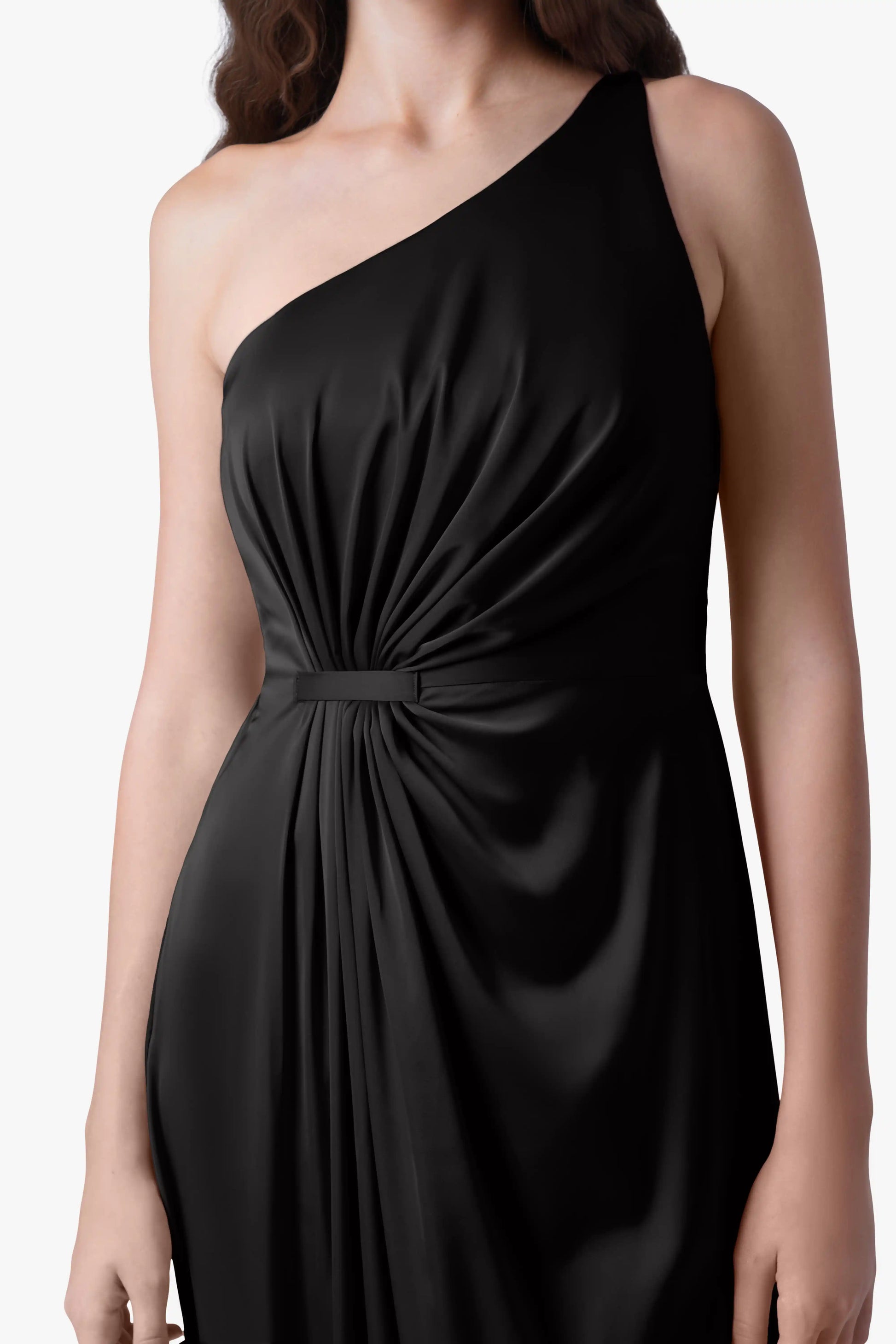 A woman models the TH&TH Greta Bridesmaid Dress in Black Satin, a sleek one-shoulder gown with Grecian side draping and gathered waist detail, standing against a plain white background.