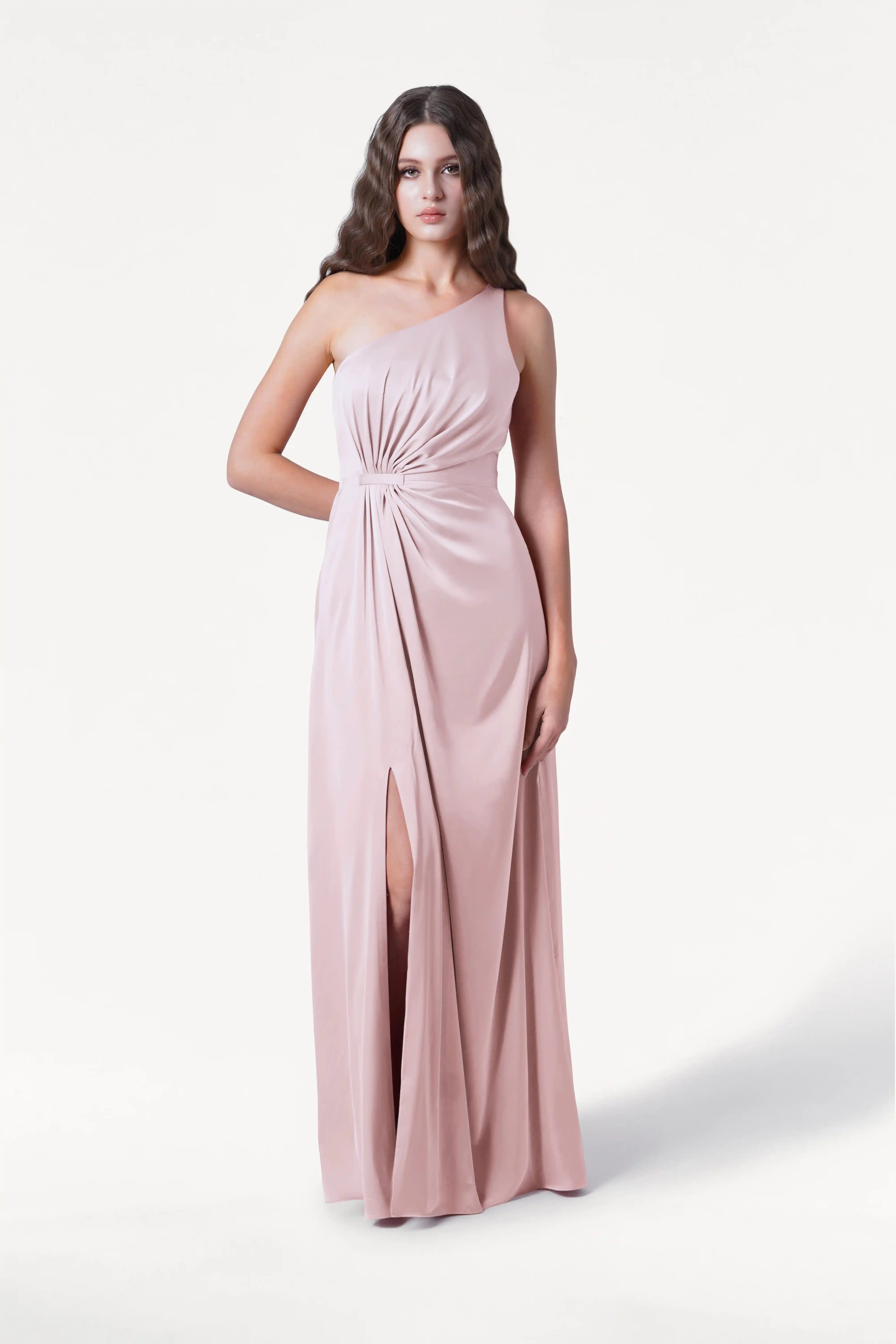 A woman wears the TH&TH Greta Bridesmaid Dress in blush pink satin, featuring a one-shoulder design, Grecian draping, gathered knot at the waist, and front slit, standing against a plain white background.
