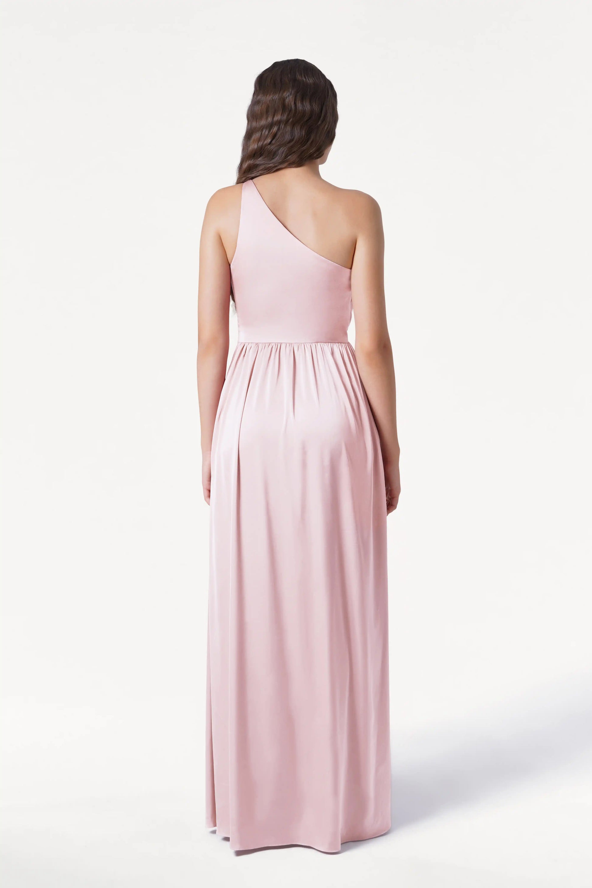 A woman with softly waved brown hair stands against a white background, wearing the TH&TH Greta Bridesmaid Dress in Blush Pink Satin—a long, one-shoulder gown with Grecian draping and a gathered waist.