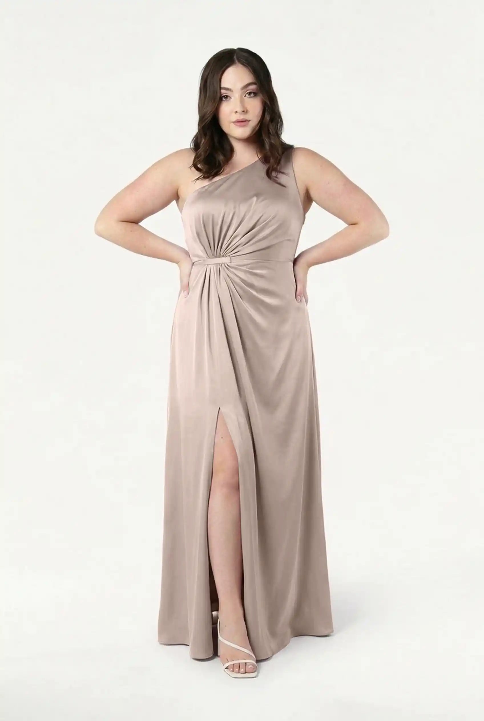 A woman faces forward in the Greta Bridesmaid Dress in Champagne Sky Satin by TH&TH, featuring a waist twist detail and front slit. She has wavy brown hair, white heeled sandals, and stands against a plain white background.