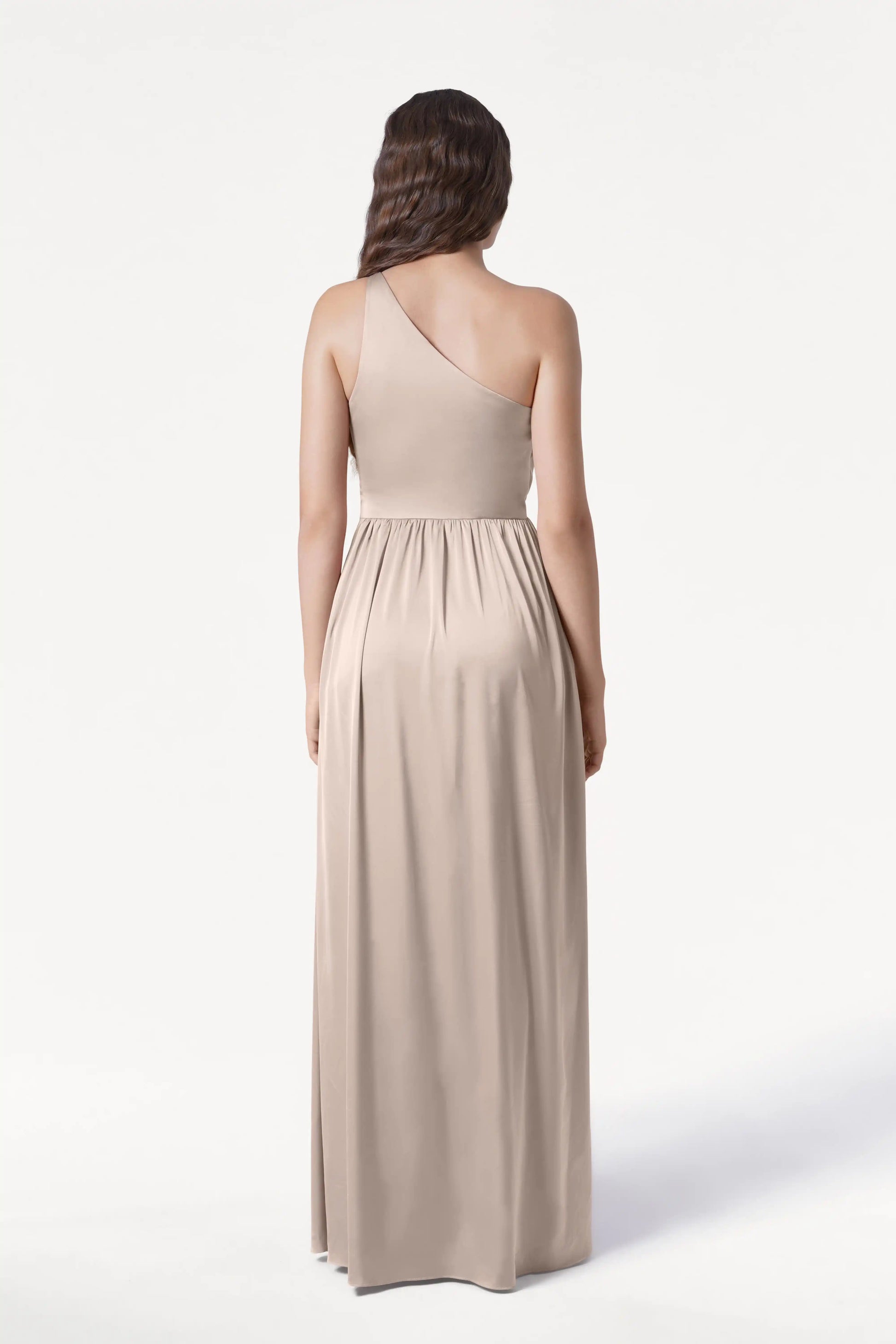A woman with long, wavy brown hair stands elegantly against a plain white background, wearing the TH&TH Greta Bridesmaid Dress in Champagne Sky Satin—a flowing, one-shoulder gown gathered at the waist.