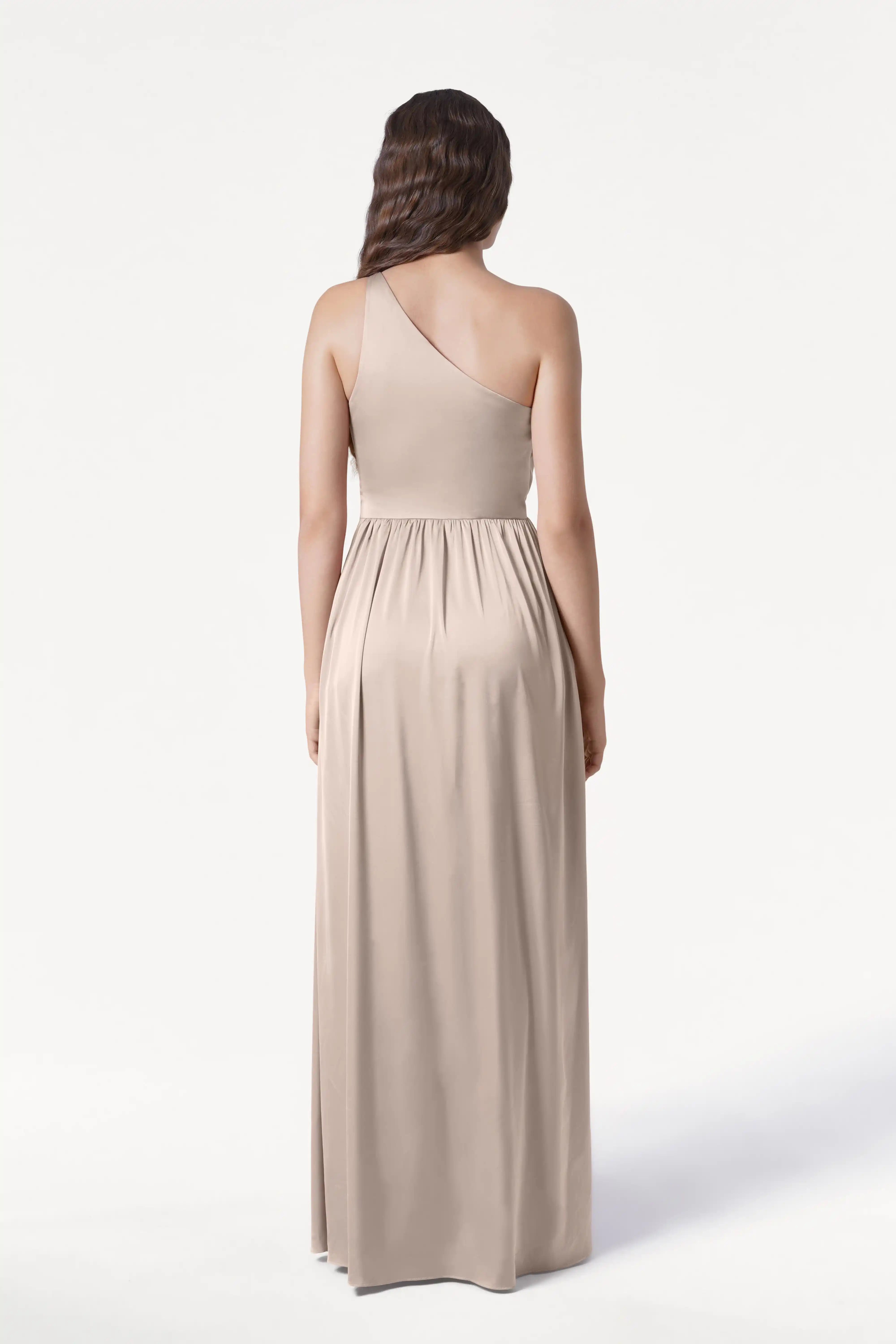A woman with long, wavy brown hair stands elegantly against a plain white background, wearing the TH&TH Greta Bridesmaid Dress in Champagne Sky Satin—a flowing, one-shoulder gown gathered at the waist.