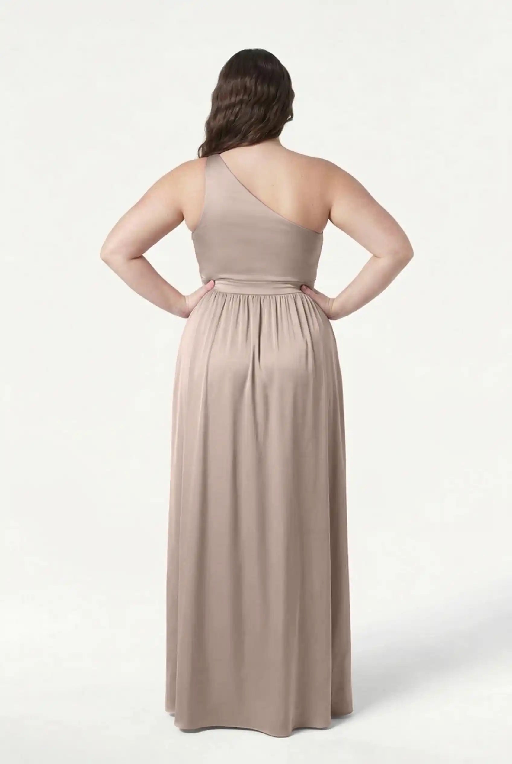 A woman stands with her back to the camera, wearing the TH&TH Greta Bridesmaid Dress in Champagne Sky Satin. She poses with bent arms against a plain white background, highlighting classic Grecian eveningwear elegance.