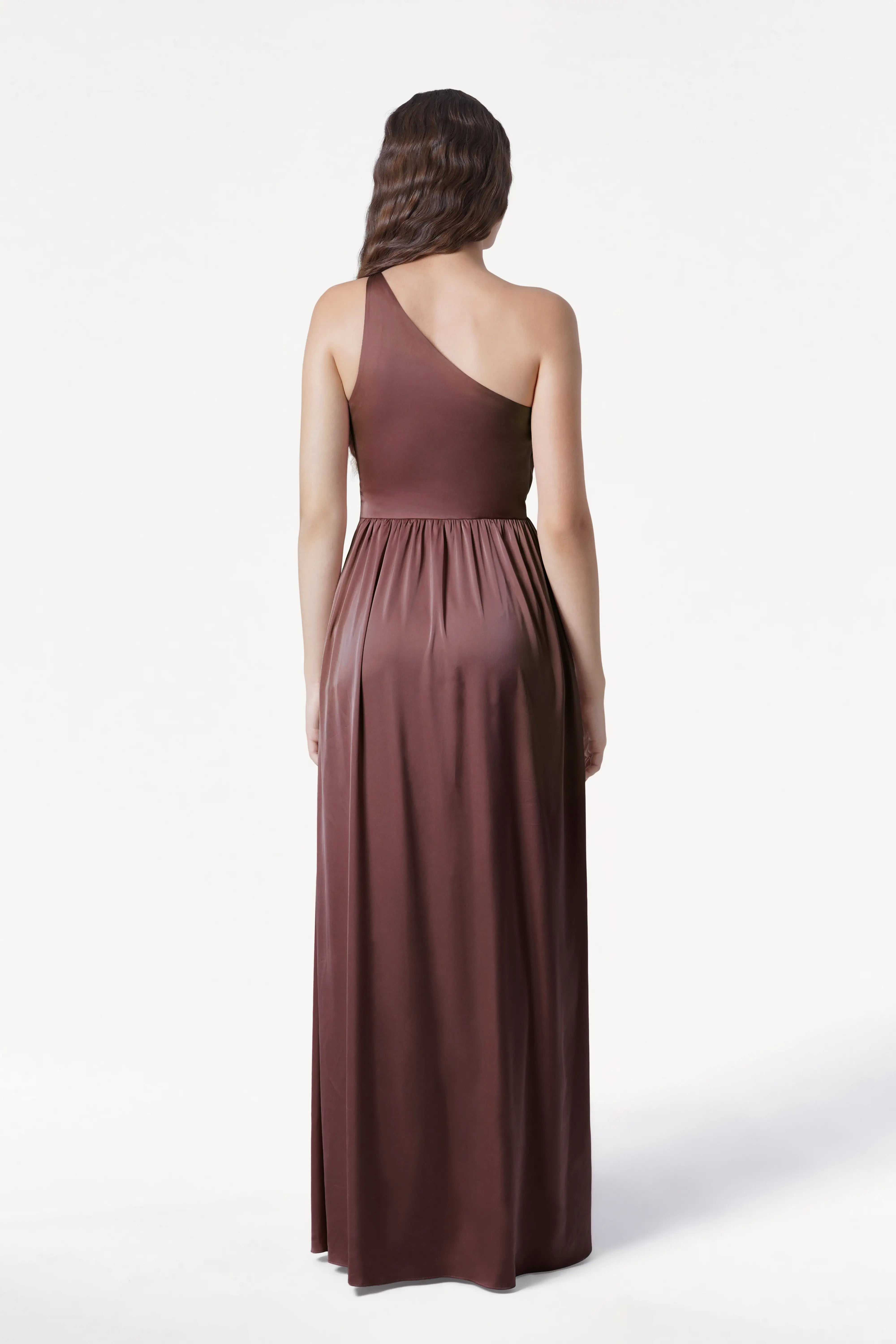 A woman with long brown hair stands facing away, wearing the TH&TH Greta Bridesmaid Dress in chocolate brown satin, a floor-length one-shoulder gown with Grecian side draping and gathered waist, against a plain white background.