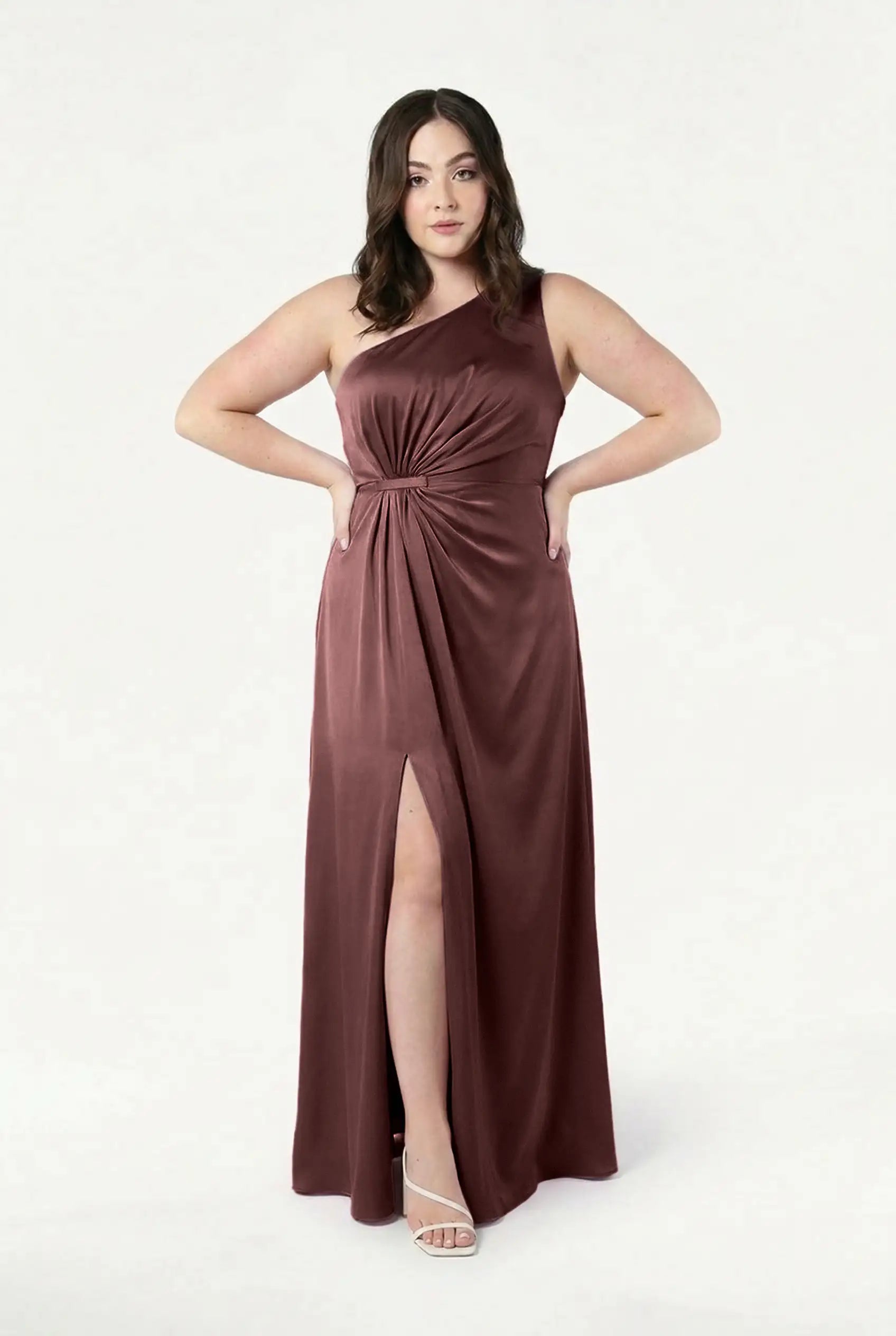 A woman stands against a white background wearing the Greta Bridesmaid Dress by TH&TH—a sleeveless, one-shoulder satin gown in chocolate brown with Grecian side draping, a knotted waist detail, and a thigh-high slit. She pairs it with white heeled sandals.
