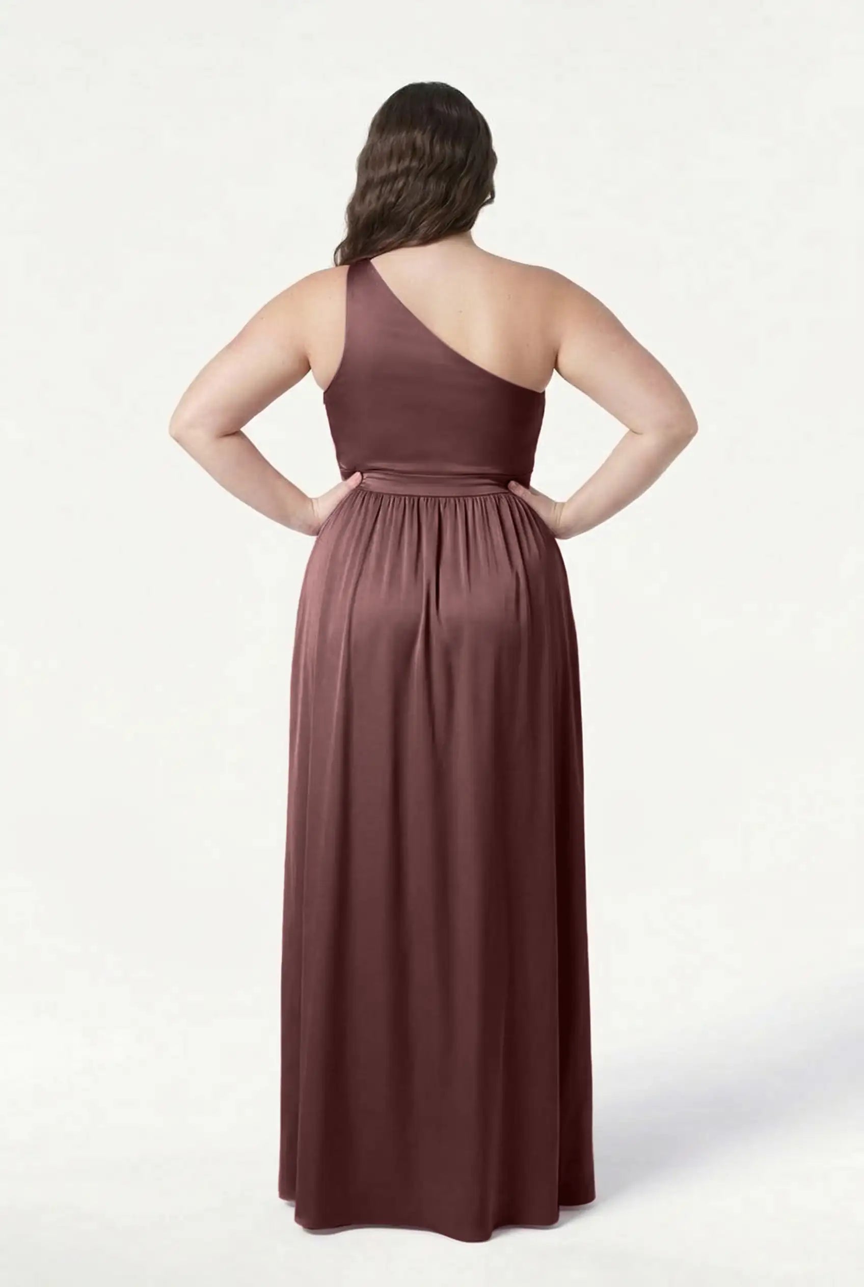 A woman stands with her back to the camera, wearing the Greta Bridesmaid Dress by TH&TH in chocolate brown satin. Her hands rest on her hips, showing off the elegant Grecian side draping against a plain white background.