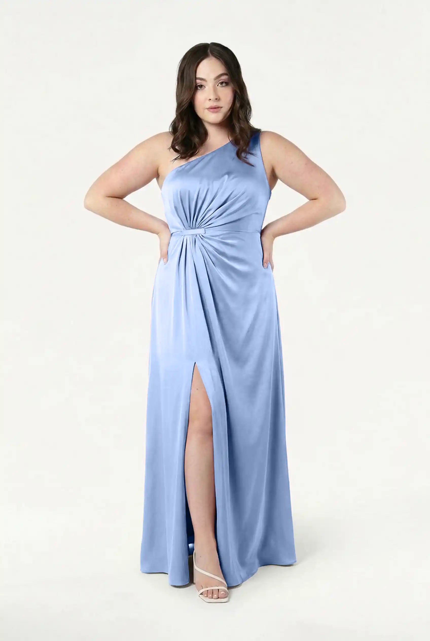 A woman wears the Greta Bridesmaid Dress in Dusty Blue Satin by TH&TH, featuring Grecian draping, a one-shoulder design, waist twist detail, and a thigh-high slit, standing with hands on hips against a plain light background.