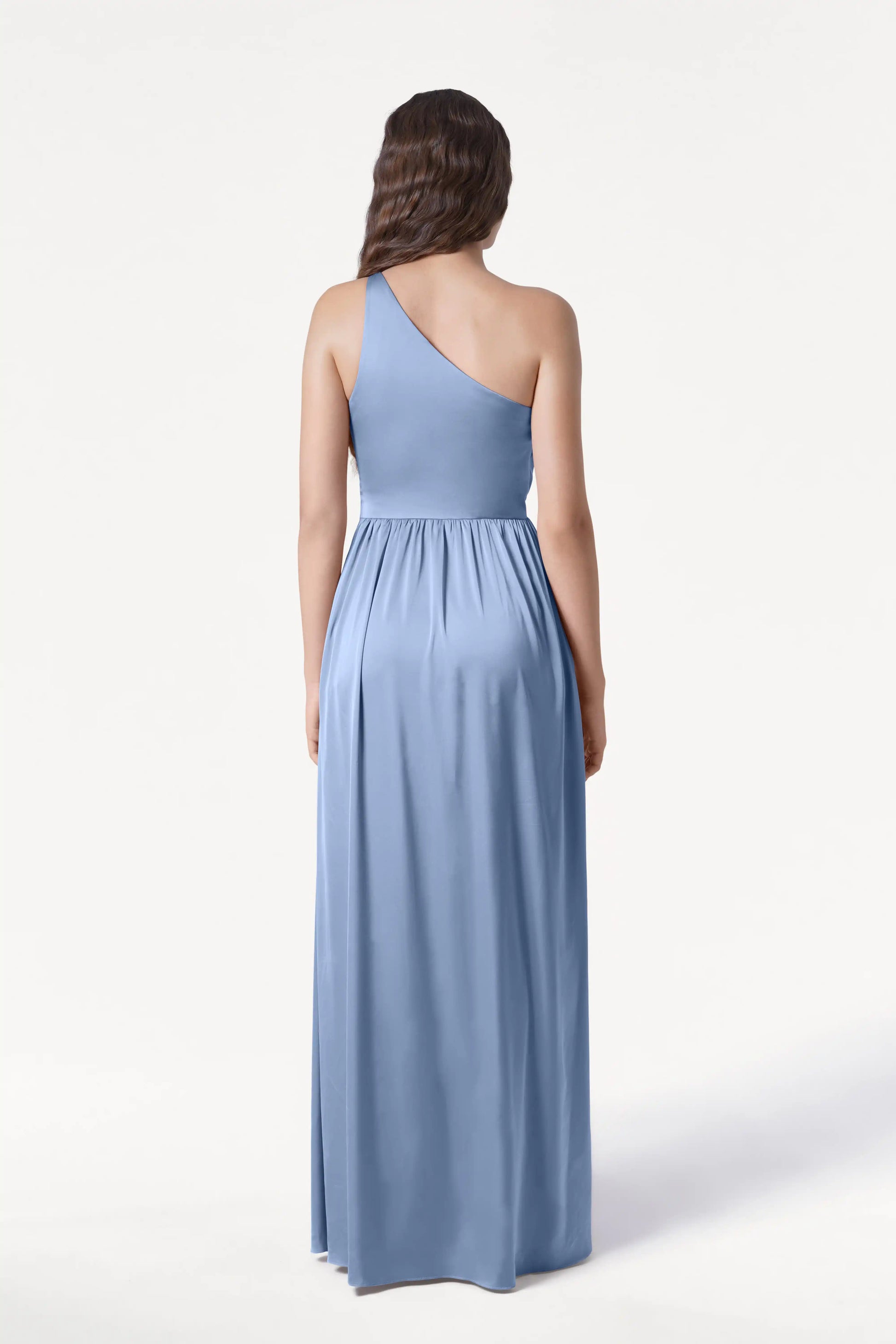 A woman with long, wavy brown hair is seen from the back wearing the TH&TH Greta Bridesmaid Dress in Dusty Blue Satin, a floor-length one-shoulder gown with elegant Grecian draping against a plain white background.