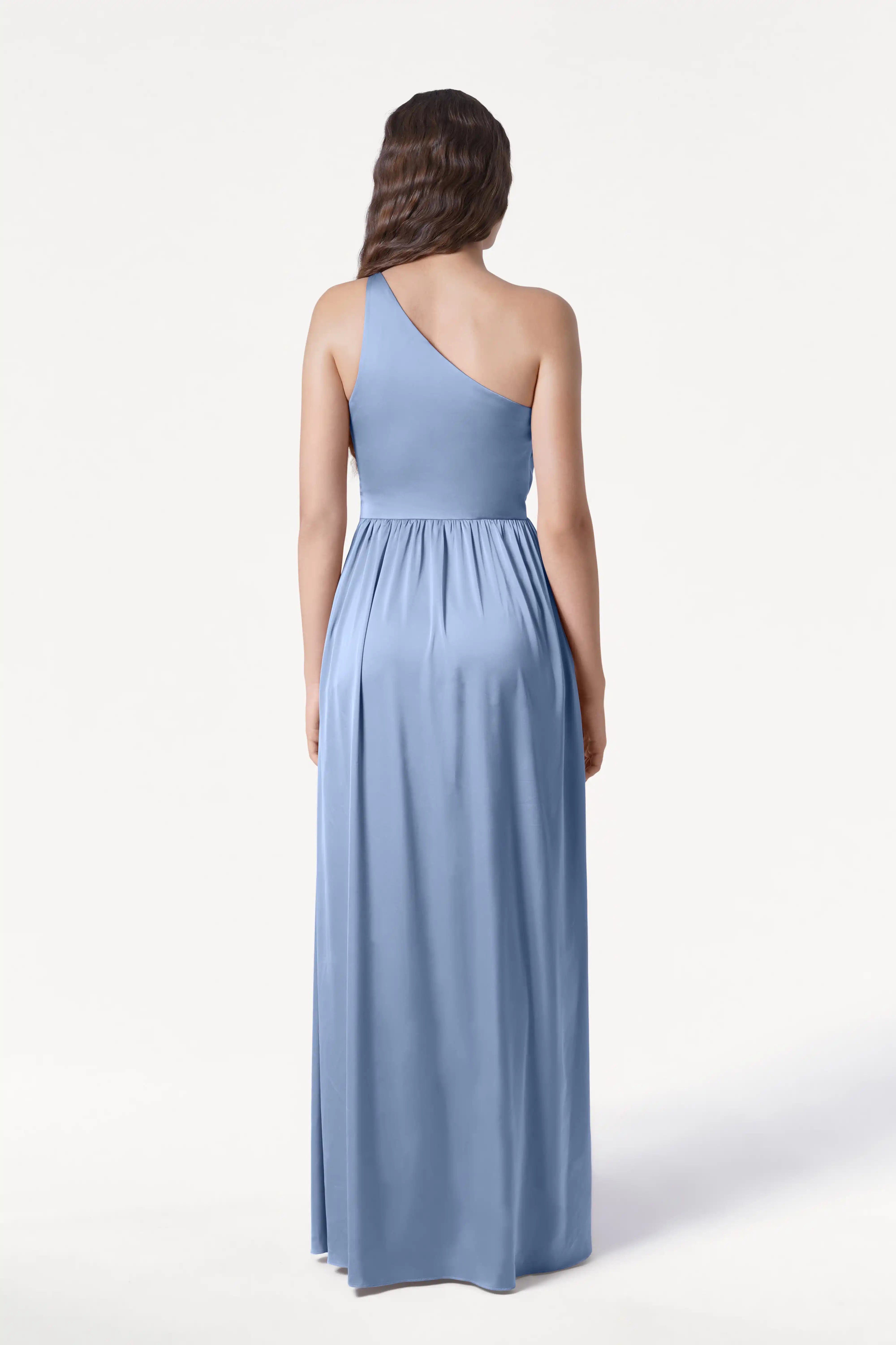 A woman with long, wavy brown hair is seen from the back wearing the TH&TH Greta Bridesmaid Dress in Dusty Blue Satin, a floor-length one-shoulder gown with elegant Grecian draping against a plain white background.