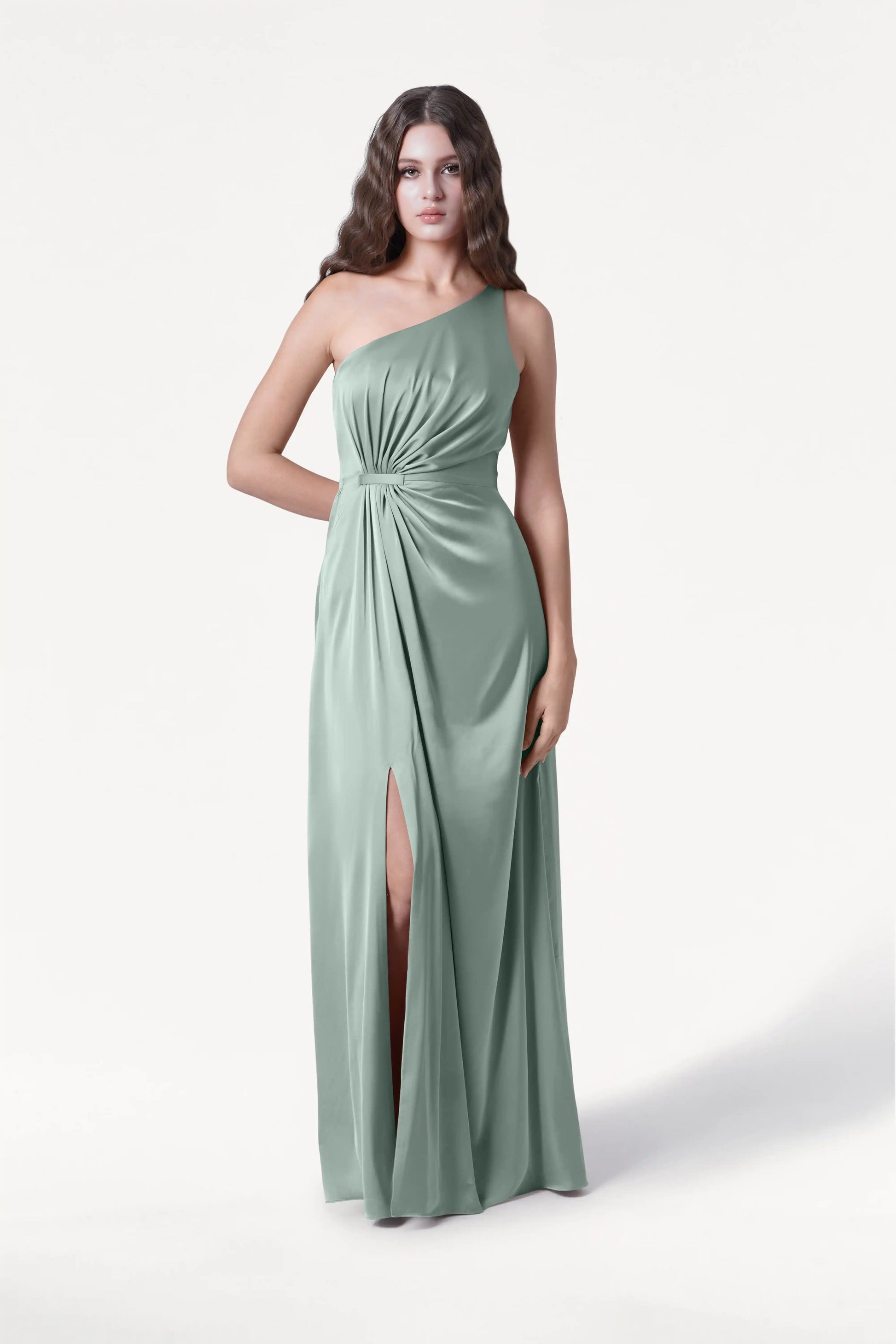 A woman with long wavy hair wears the TH&TH Greta Bridesmaid Dress in Dusty Eucalyptus Satin, a one-shoulder gown with Grecian draping, gathered waist, and thigh-high slit, standing against a plain white background.