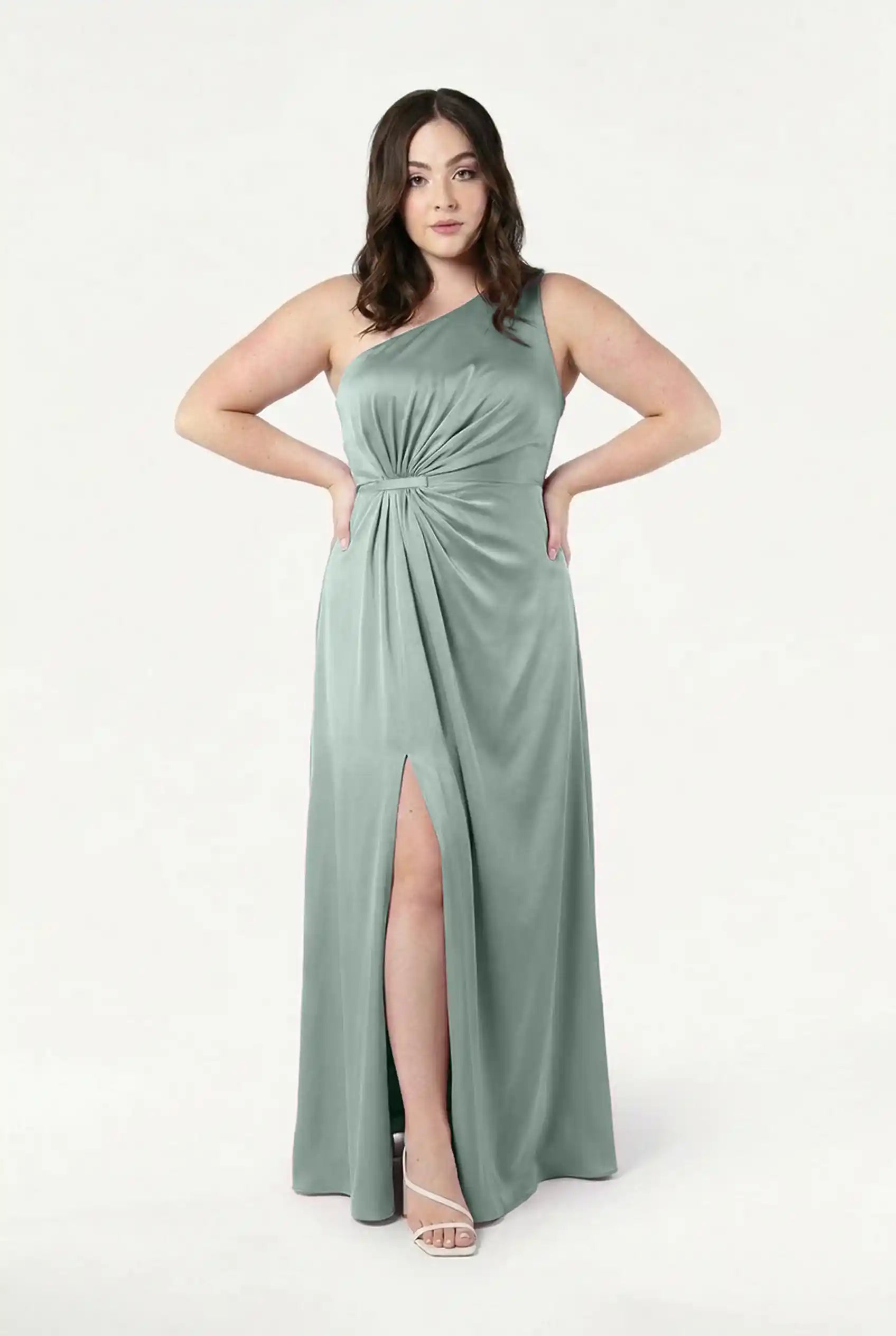 A woman wears the TH&TH Greta Bridesmaid Dress in Dusty Eucalyptus Satin, featuring Grecian draping, a waist knot detail, one-shoulder design, and thigh-high slit as she poses confidently against a plain white background.