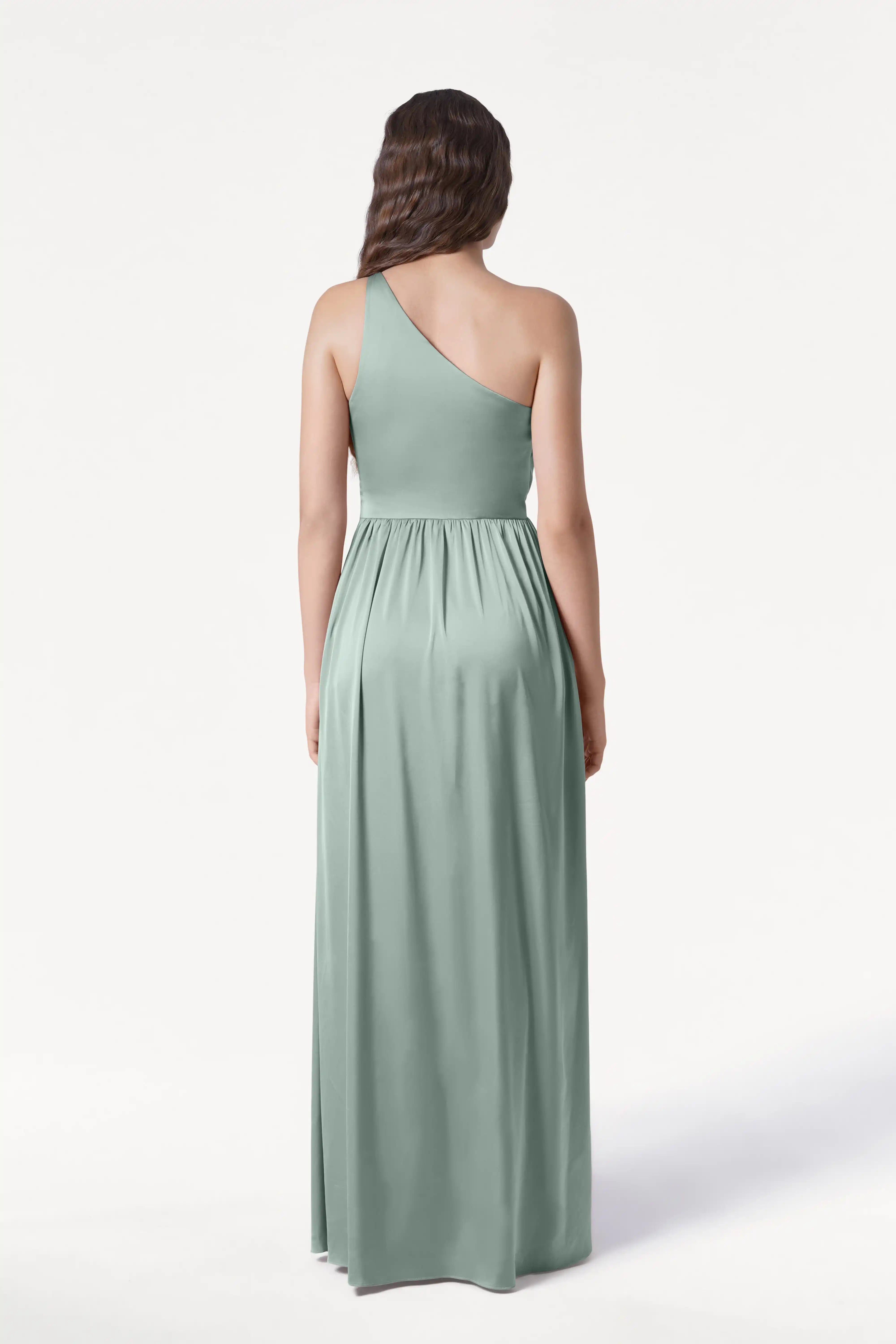 A woman faces away, wearing the TH&TH Greta Bridesmaid Dress in Dusty Eucalyptus Satin—a floor-length gown with Grecian draping and a gathered waist—against a plain white background.