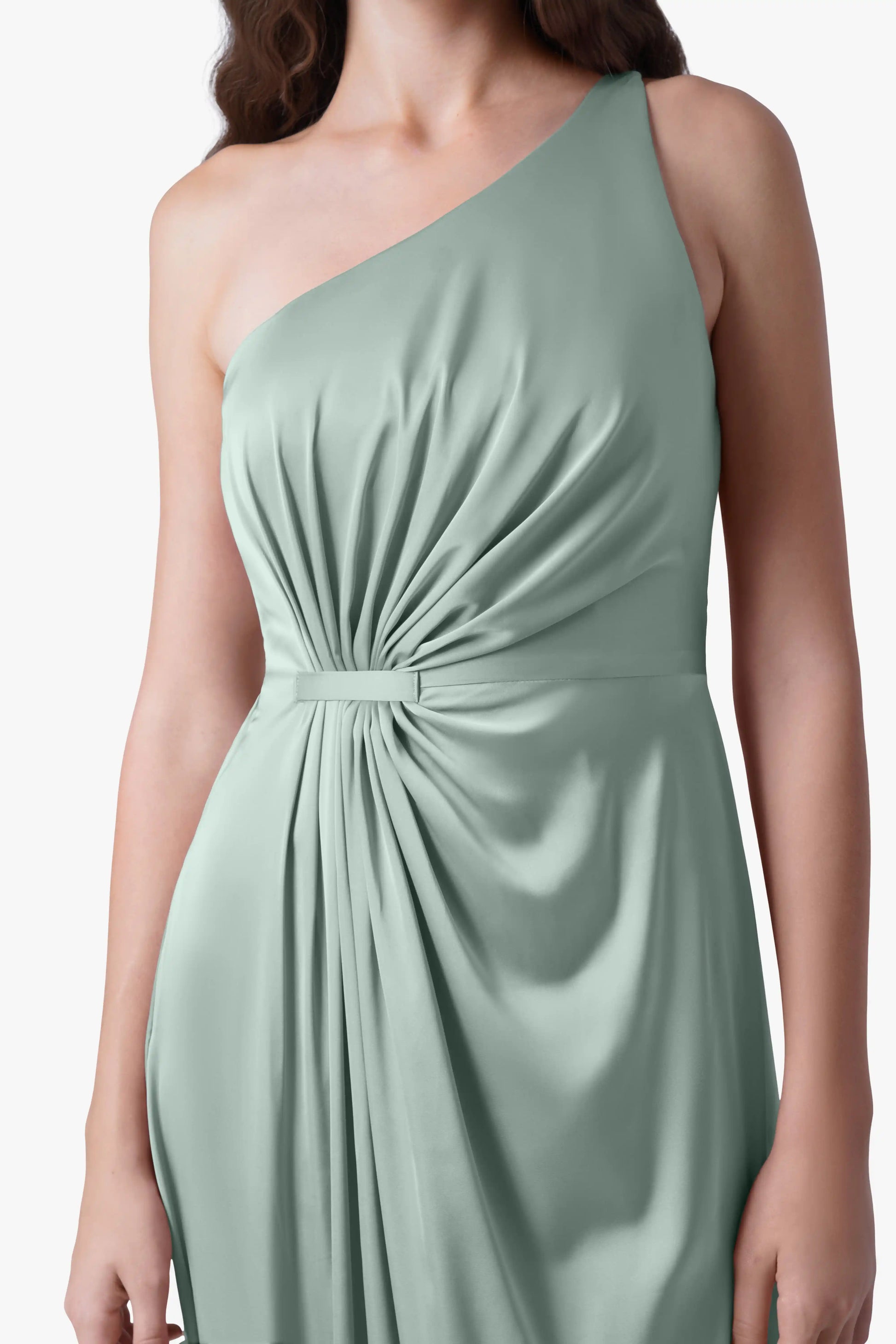 A woman wears the TH&TH Greta Bridesmaid Dress in Dusty Eucalyptus Satin, featuring a sleeveless one-shoulder design, Grecian draping, ruched waist, and a belt accent. The soft, flowing fabric stands out; her head is not visible.