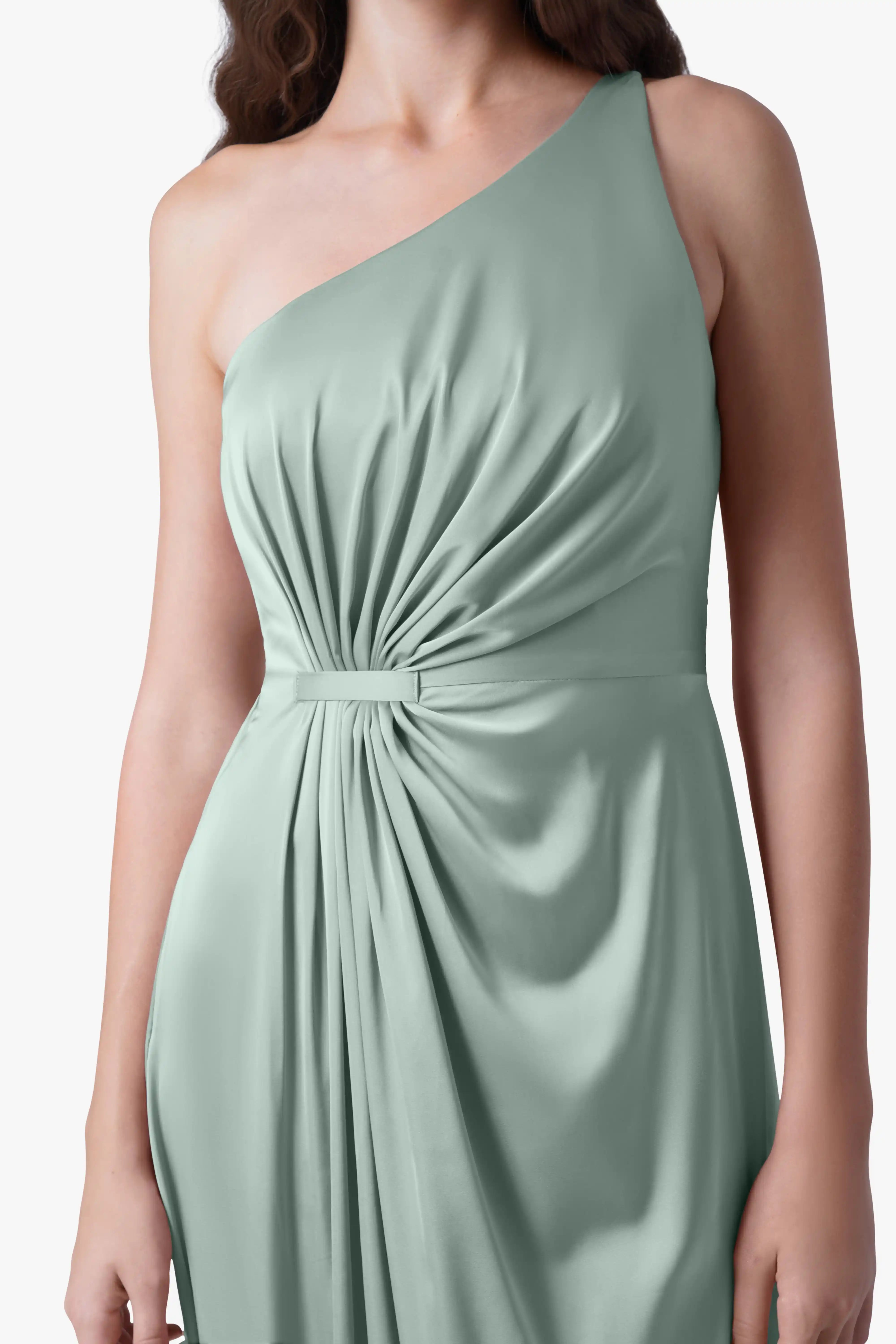 A woman wears the TH&TH Greta Bridesmaid Dress in Dusty Eucalyptus Satin, featuring a sleeveless one-shoulder design, Grecian draping, ruched waist, and a belt accent. The soft, flowing fabric stands out; her head is not visible.