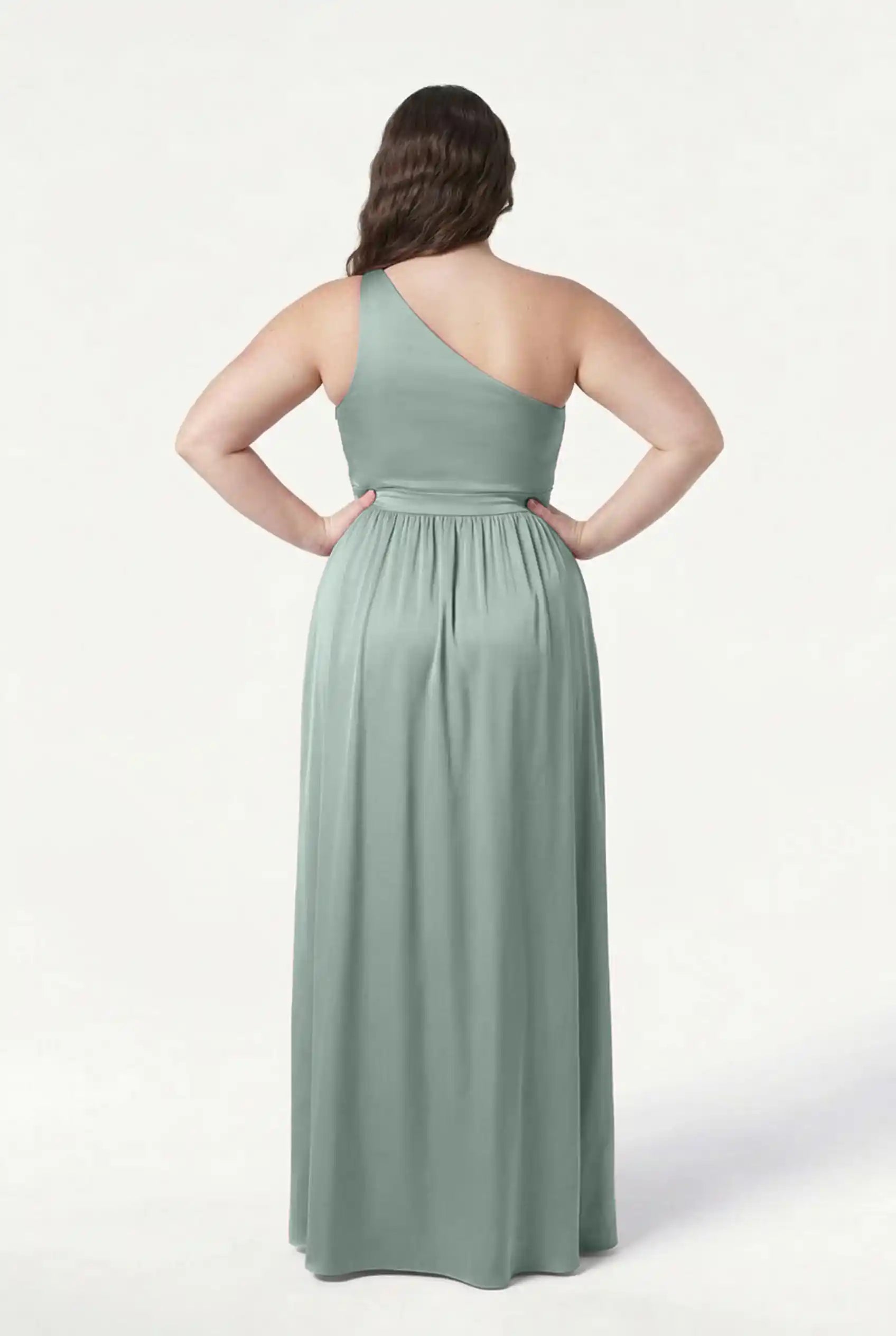 A woman stands with her back to the camera, hands on hips, wearing the TH&TH Greta Bridesmaid Dress in Dusty Eucalyptus Satin. The one-shoulder, floor-length gown features a gathered waist and flowing skirt against a plain white background.