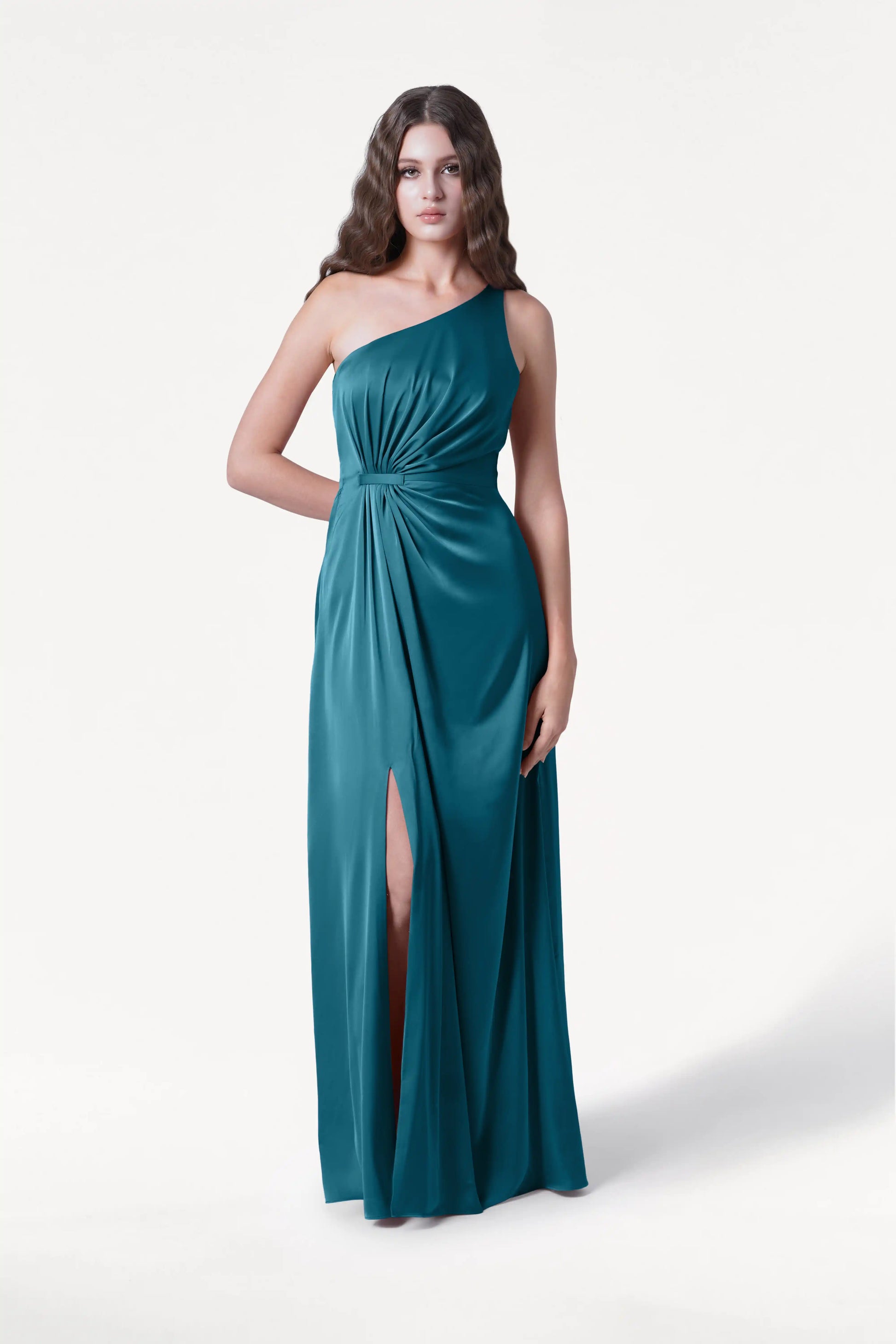 A woman with long wavy brown hair wears the TH&TH Greta Bridesmaid Dress in Emerald Dawn Satin, a floor-length one-shoulder gown with a gathered waist and thigh-high slit, standing against a plain white background.