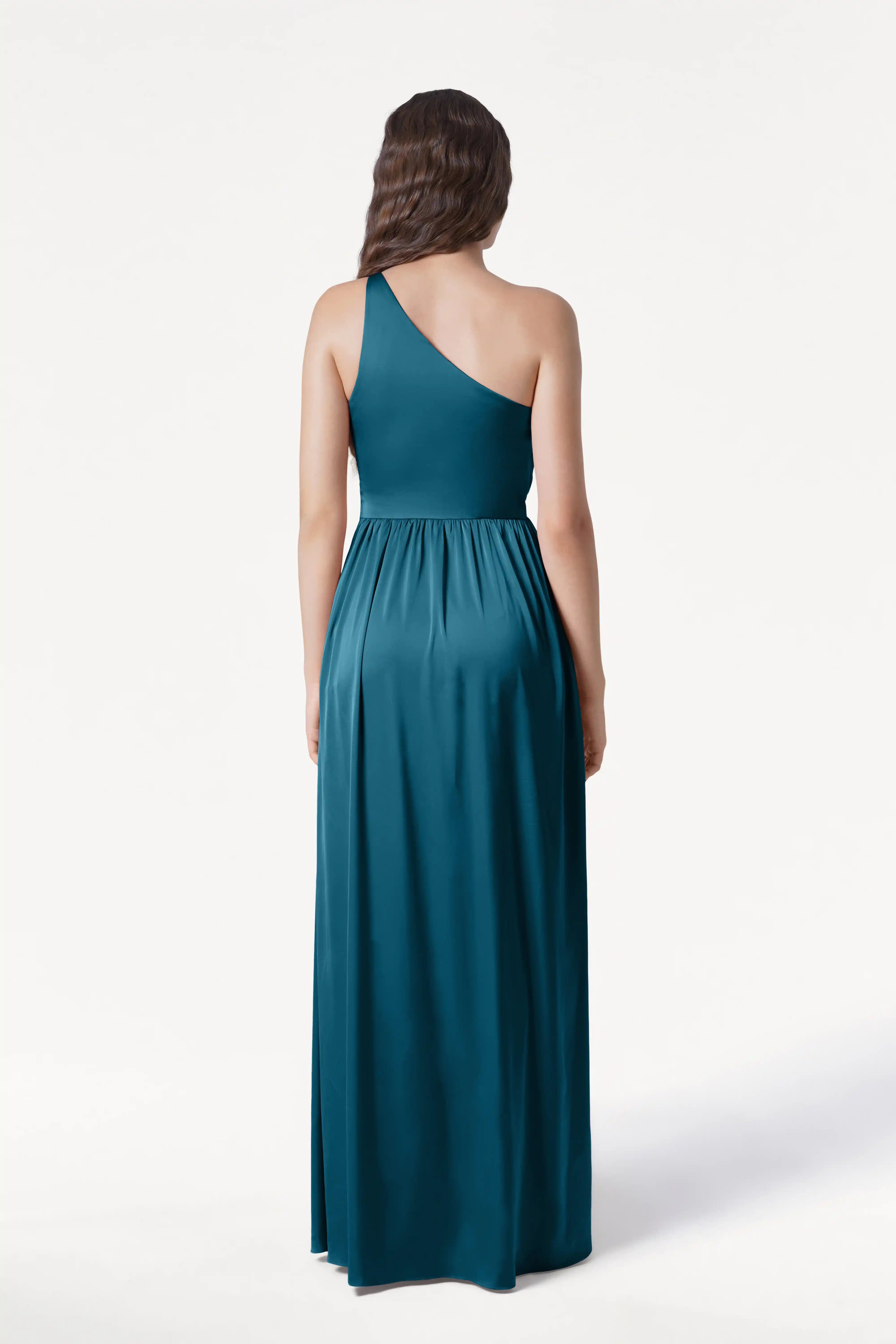 A woman with long, wavy brown hair stands with her back to the camera, wearing the TH&TH Greta Bridesmaid Dress in Emerald Dawn Satin against a plain white background.