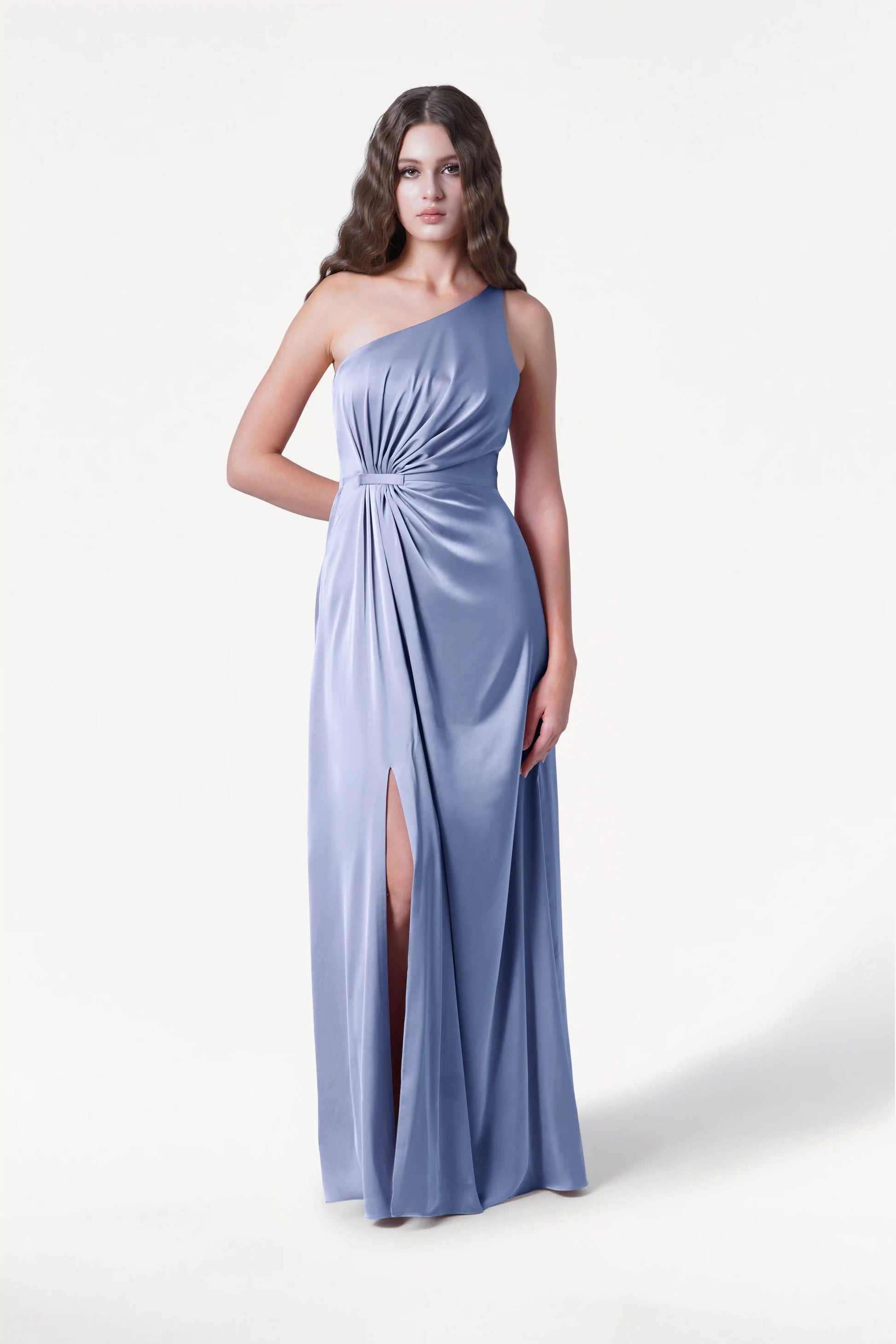 A woman models the TH&TH Greta Bridesmaid Dress in French Blue Satin—a one-shoulder gown with a gathered knot at the waist and high skirt slit—against a plain white background, highlighting its elegant eveningwear style.