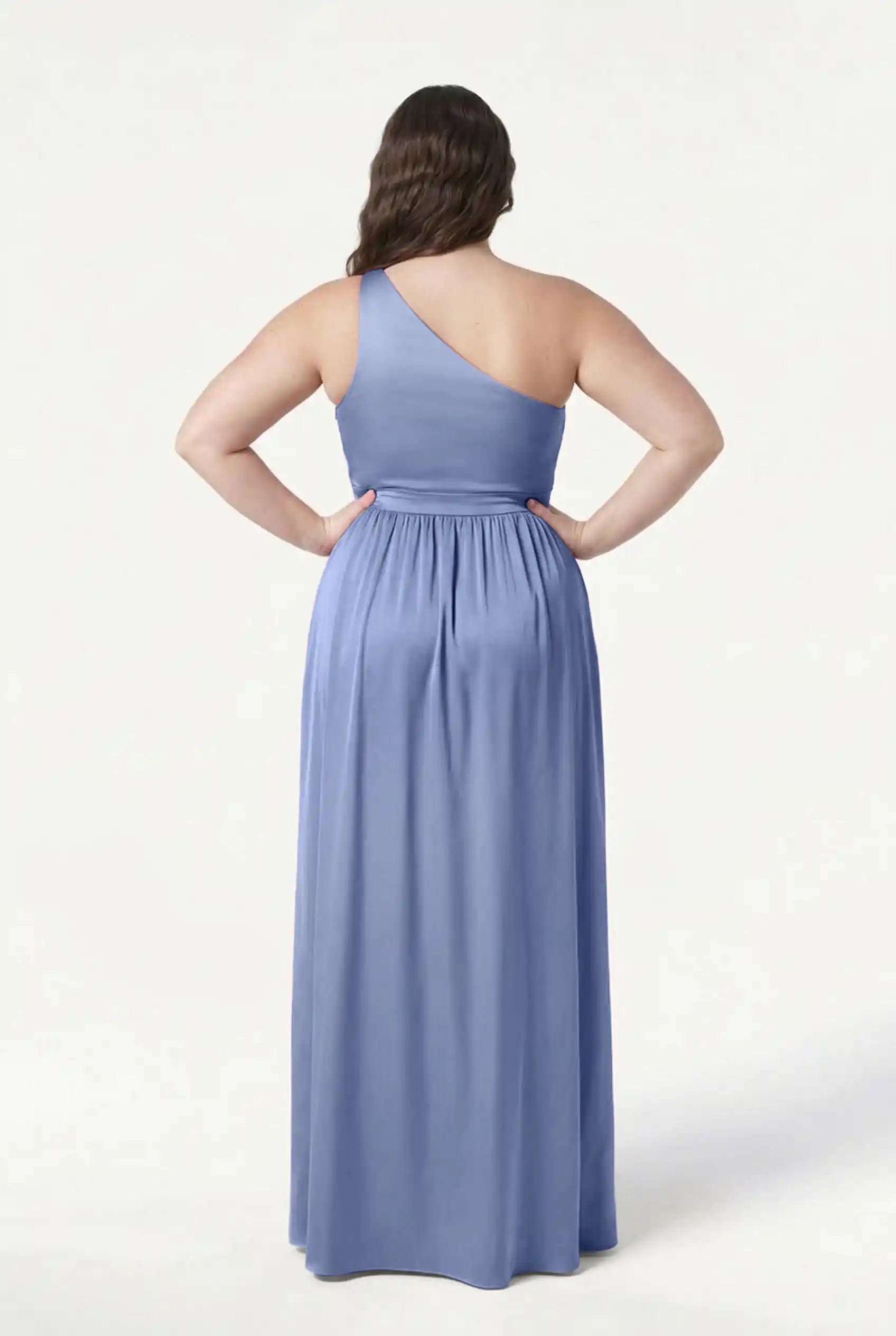 A woman stands facing away, hands on hips, wearing the TH&TH Greta Bridesmaid Dress in French Blue Satin. This elegant gown features a gathered waist and one-shoulder design, set against a plain white background.