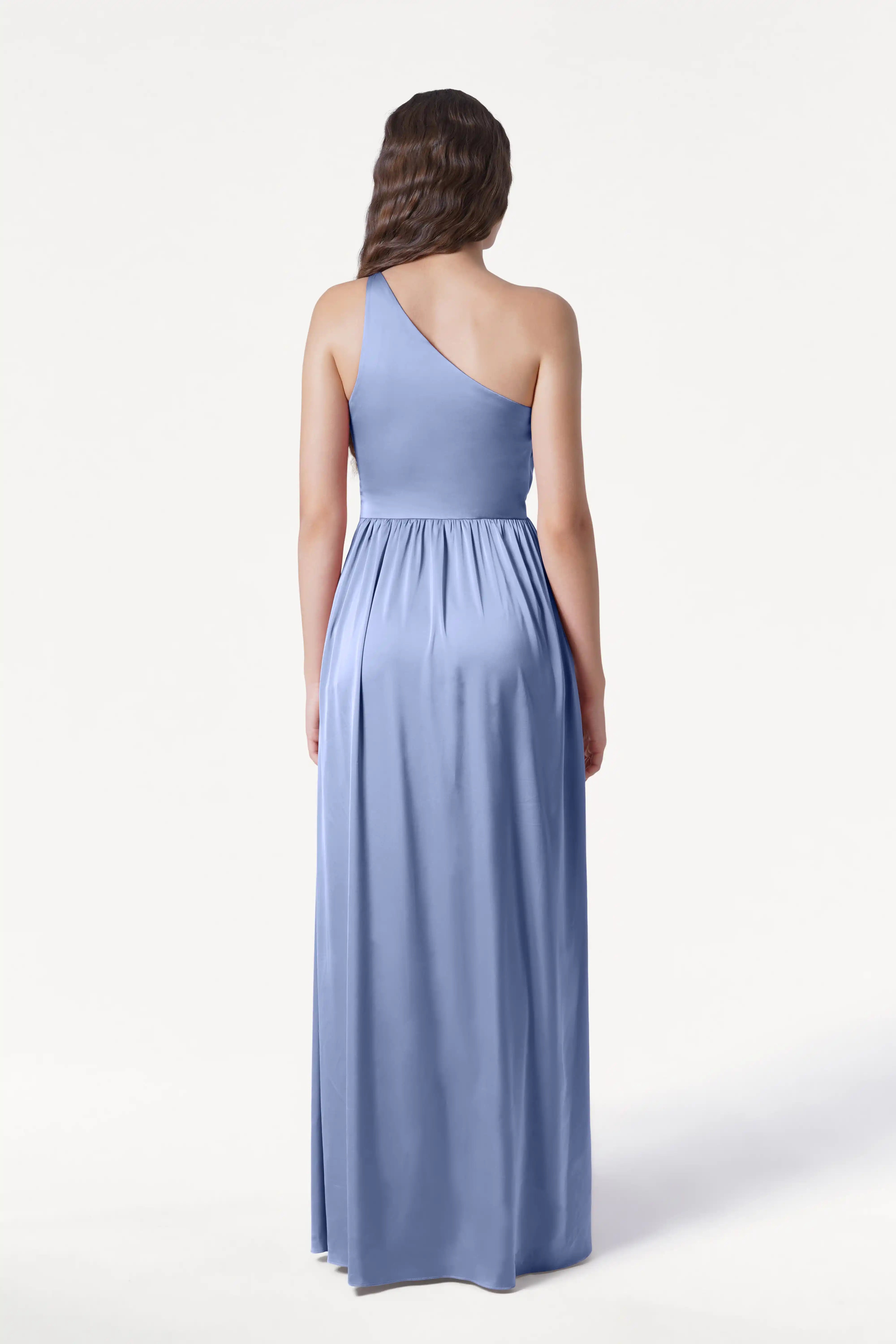 A woman faces away, wearing the TH&TH Greta Bridesmaid Dress in French Blue Satin. The floor-length gown features a gathered waist and one-shoulder design, with her long hair flowing in waves against a plain white background.
