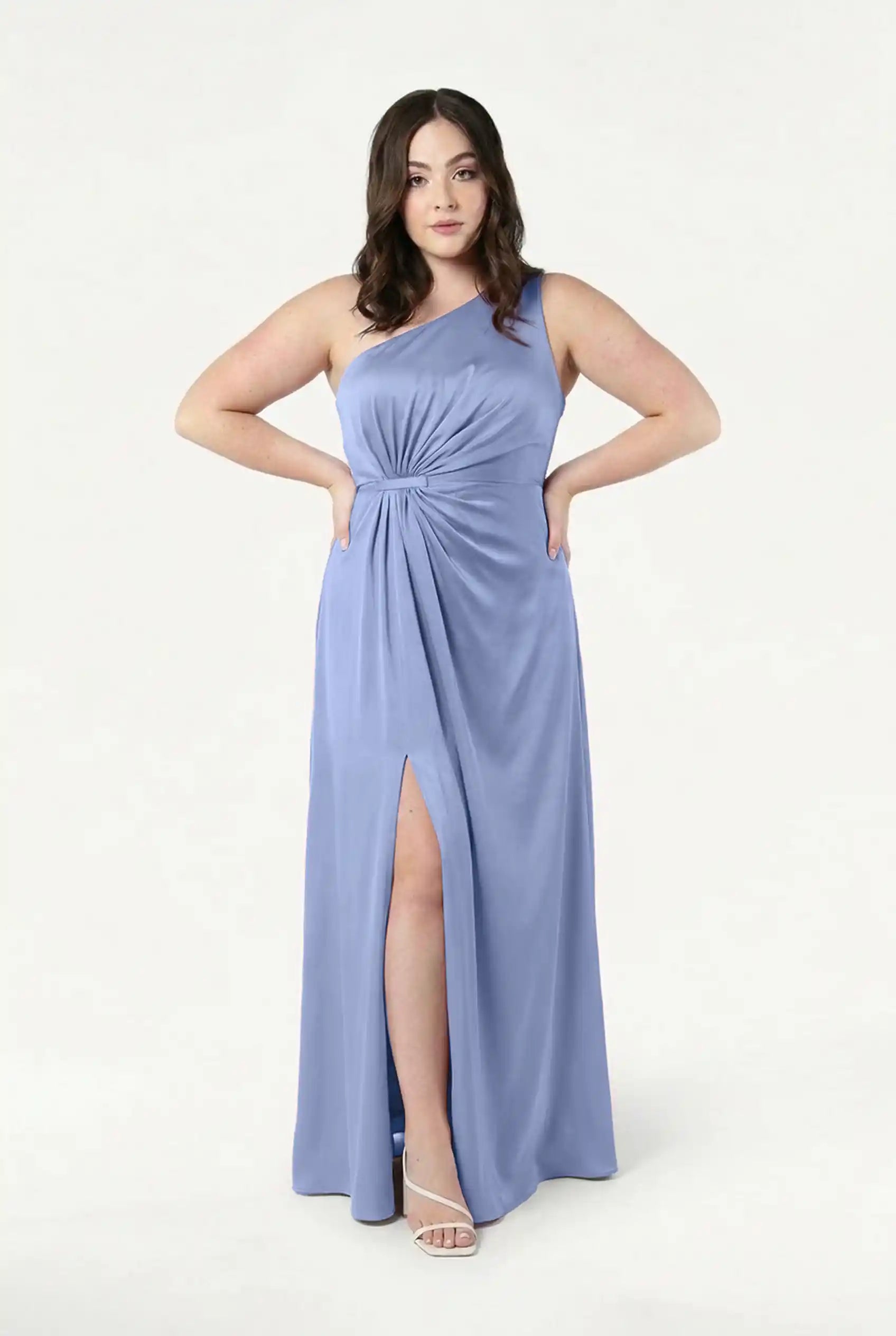 A woman stands confidently in the TH&TH Greta Bridesmaid Dress in French Blue Satin, featuring a one-shoulder design, a knotted waist detail, and a front slit. She completes the elegant look with white strappy heels.
