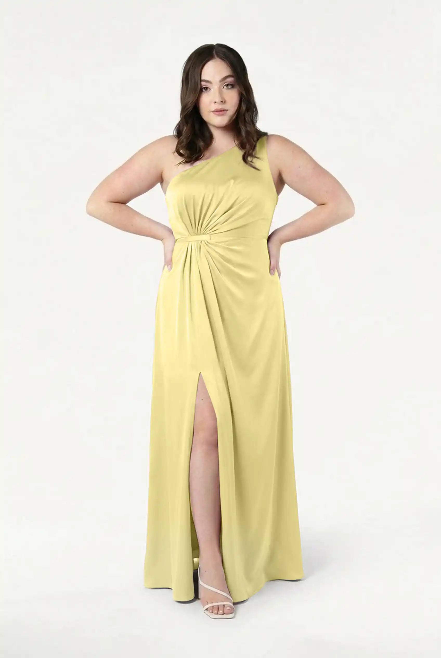 A woman models the TH&TH Greta Bridesmaid Dress in Limoncello Satin, a light yellow, one-shoulder gown with Grecian draping, gathered waist, thigh-high slit, and white strappy heels against a plain white background.