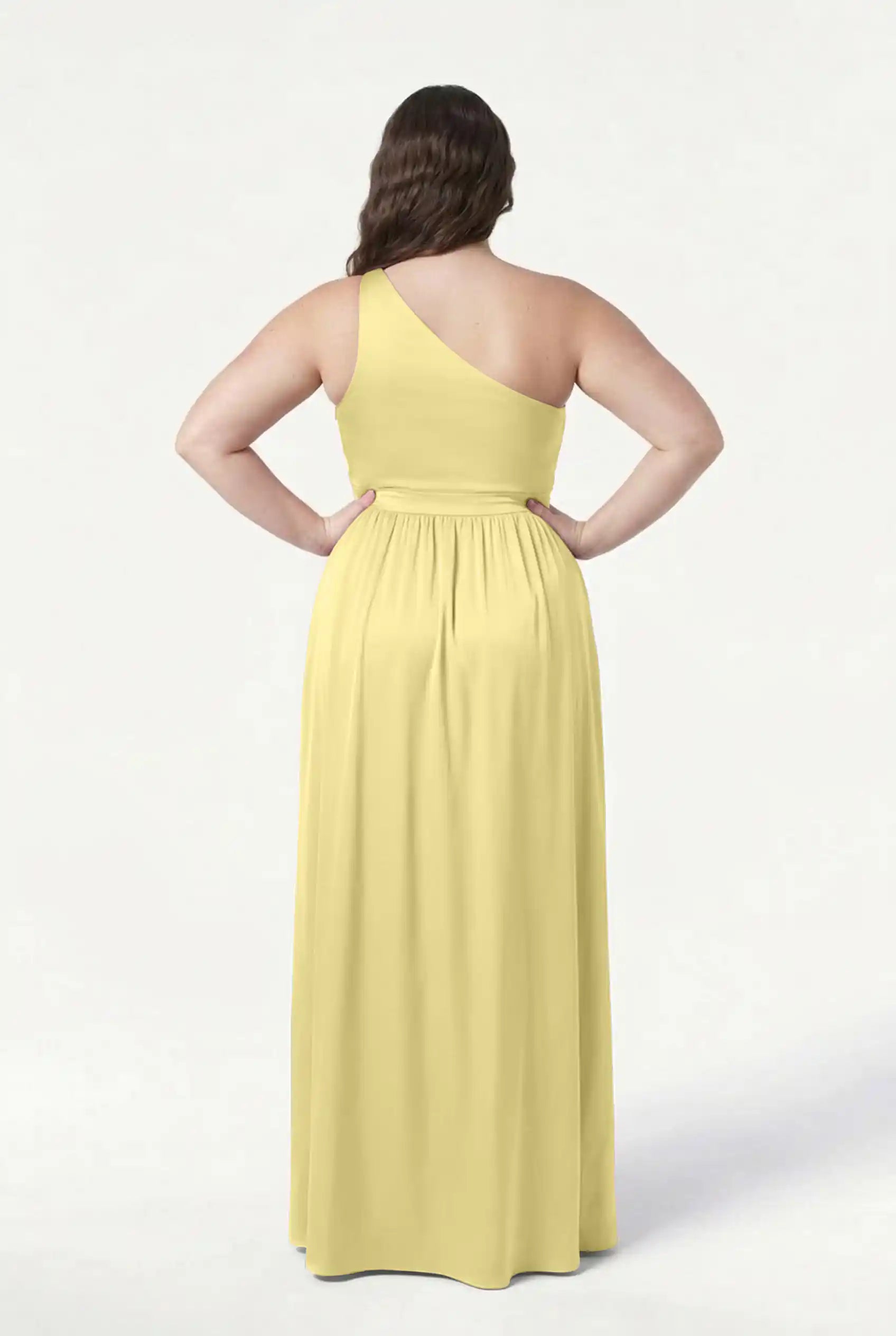 A person with long brown hair faces away, wearing the floor-length TH&TH Greta Bridesmaid Dress in Limoncello Satin, featuring Grecian draping and a gathered waist, against a plain white background.