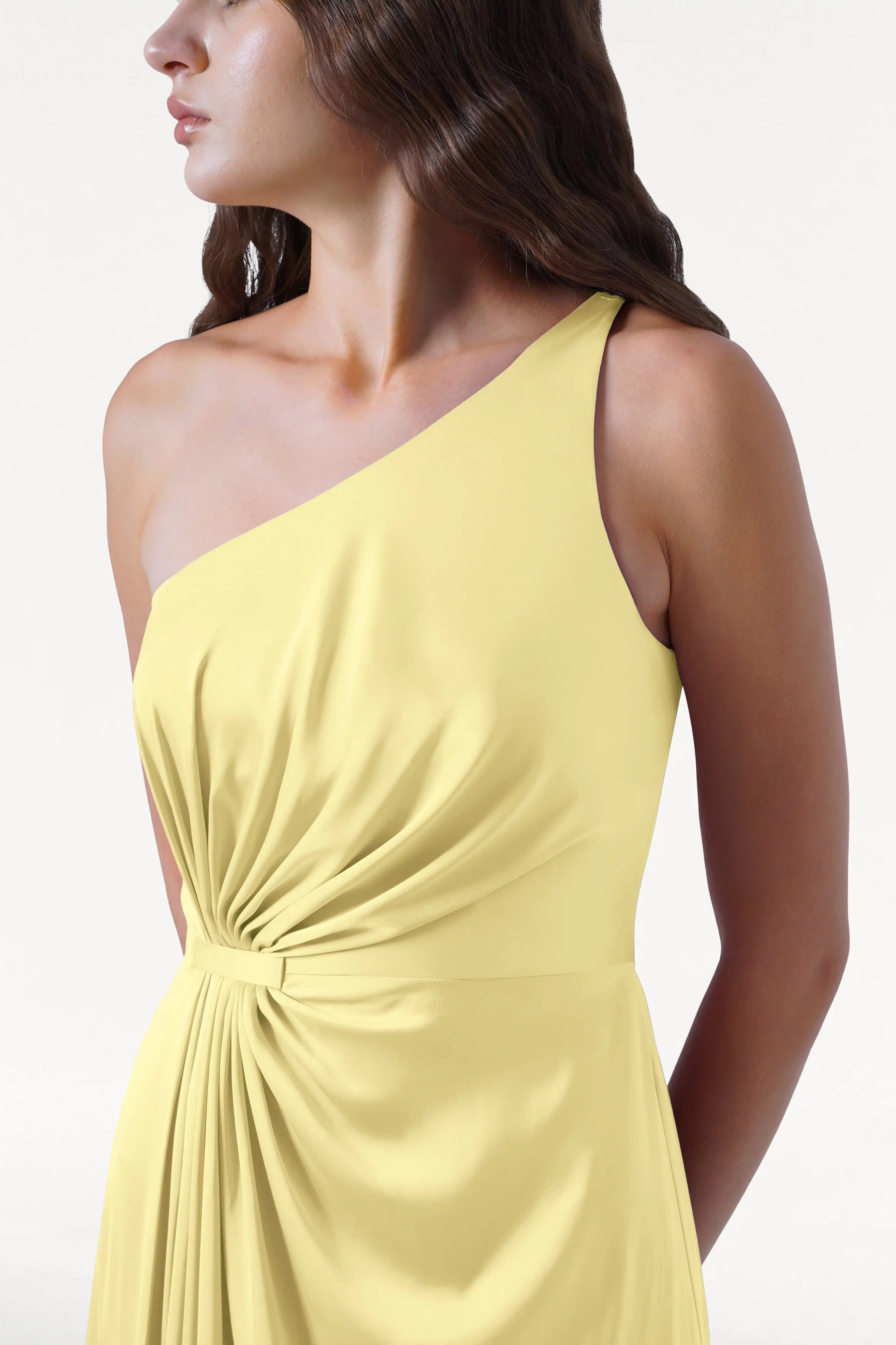 A woman with wavy brown hair wears the TH&TH Greta Bridesmaid Dress in Limoncello Satin, a pale yellow, one-shoulder gown with Grecian draping and a knotted waist, standing slightly turned to the side against a plain white background.