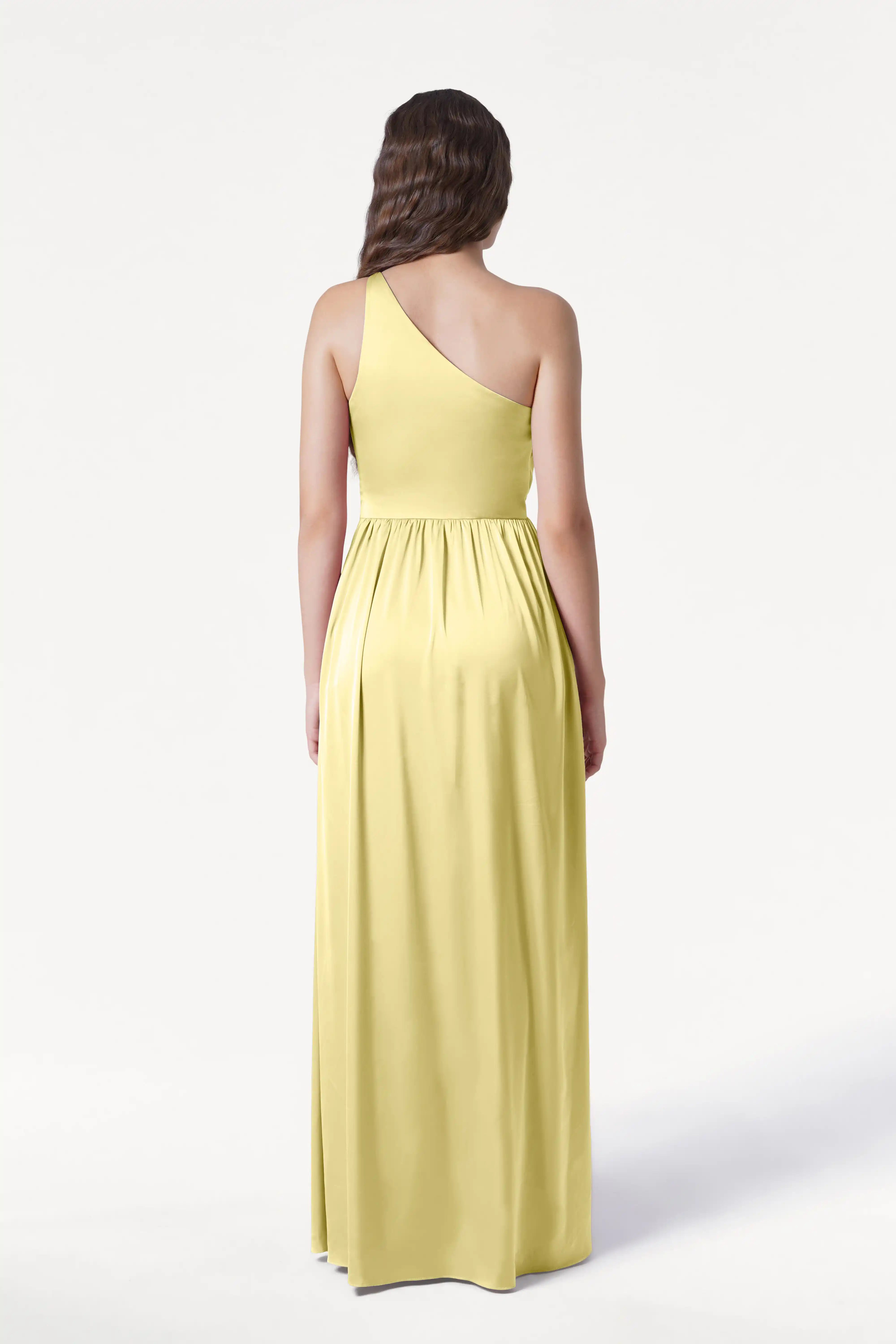A woman with wavy brown hair stands facing away, wearing the TH&TH Greta Bridesmaid Dress in Limoncello Satin—a long gown with Grecian draping and a gathered waist—set against a plain white background.