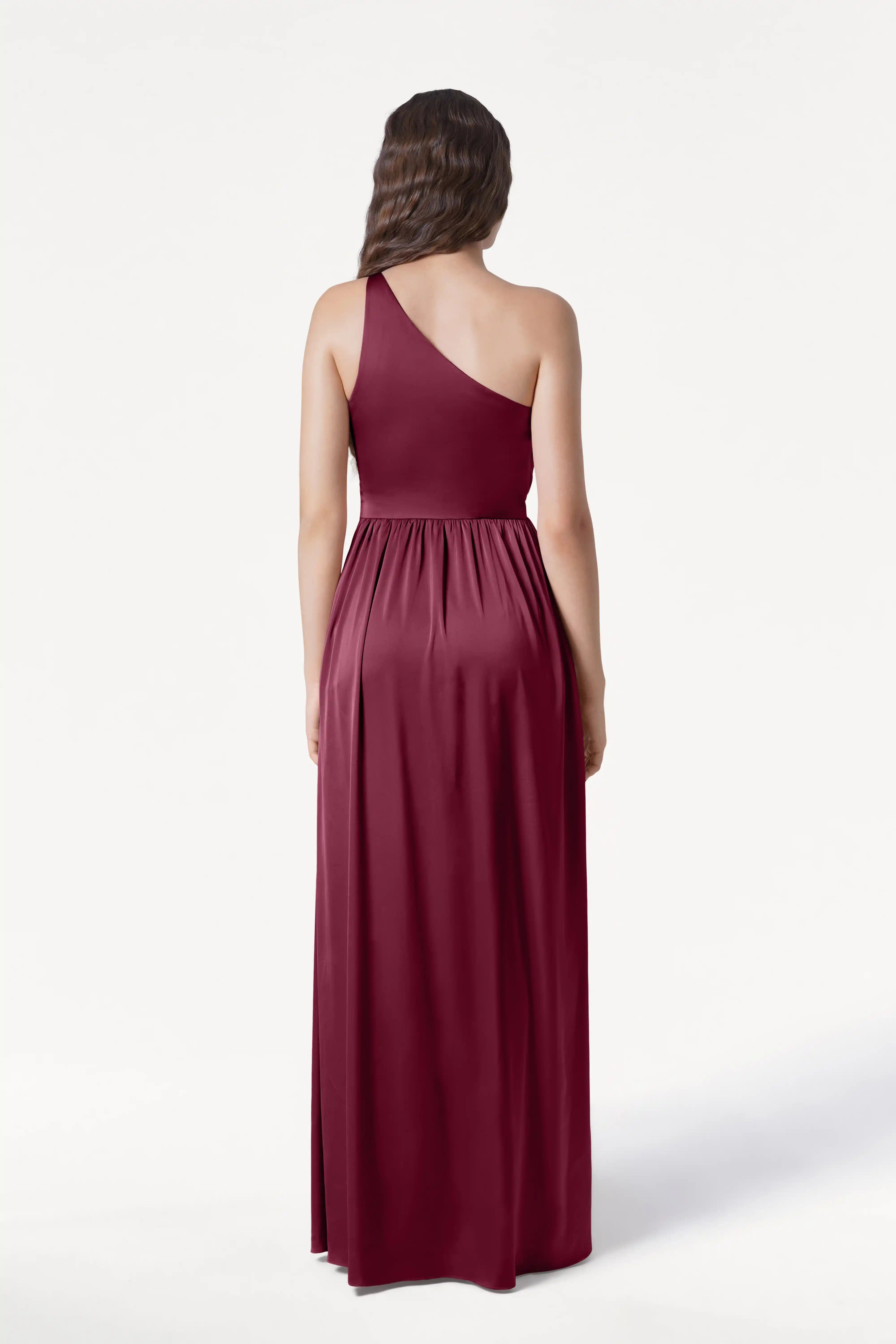 A woman with long wavy brown hair models the TH&TH Greta Bridesmaid Dress in Merlot Satin, a floor-length, sleeveless one-shoulder evening gown, shown from the back against a plain white background.