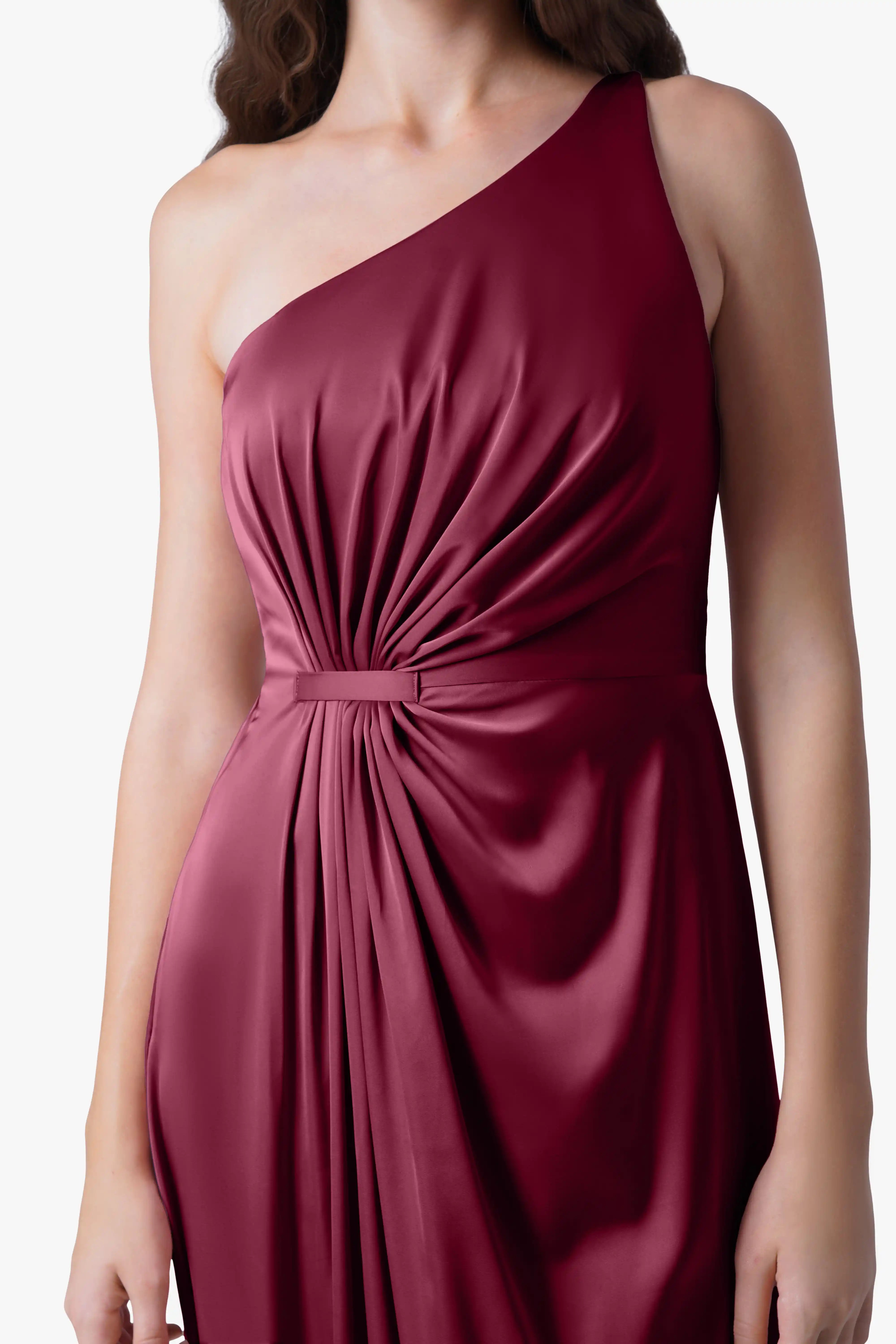 A woman models the TH&TH Greta Bridesmaid Dress in Merlot Satin, a sleeveless one-shoulder gown with gathered draping at the waist, inspired by Grecian eveningwear, standing against a plain white background with her face unseen.