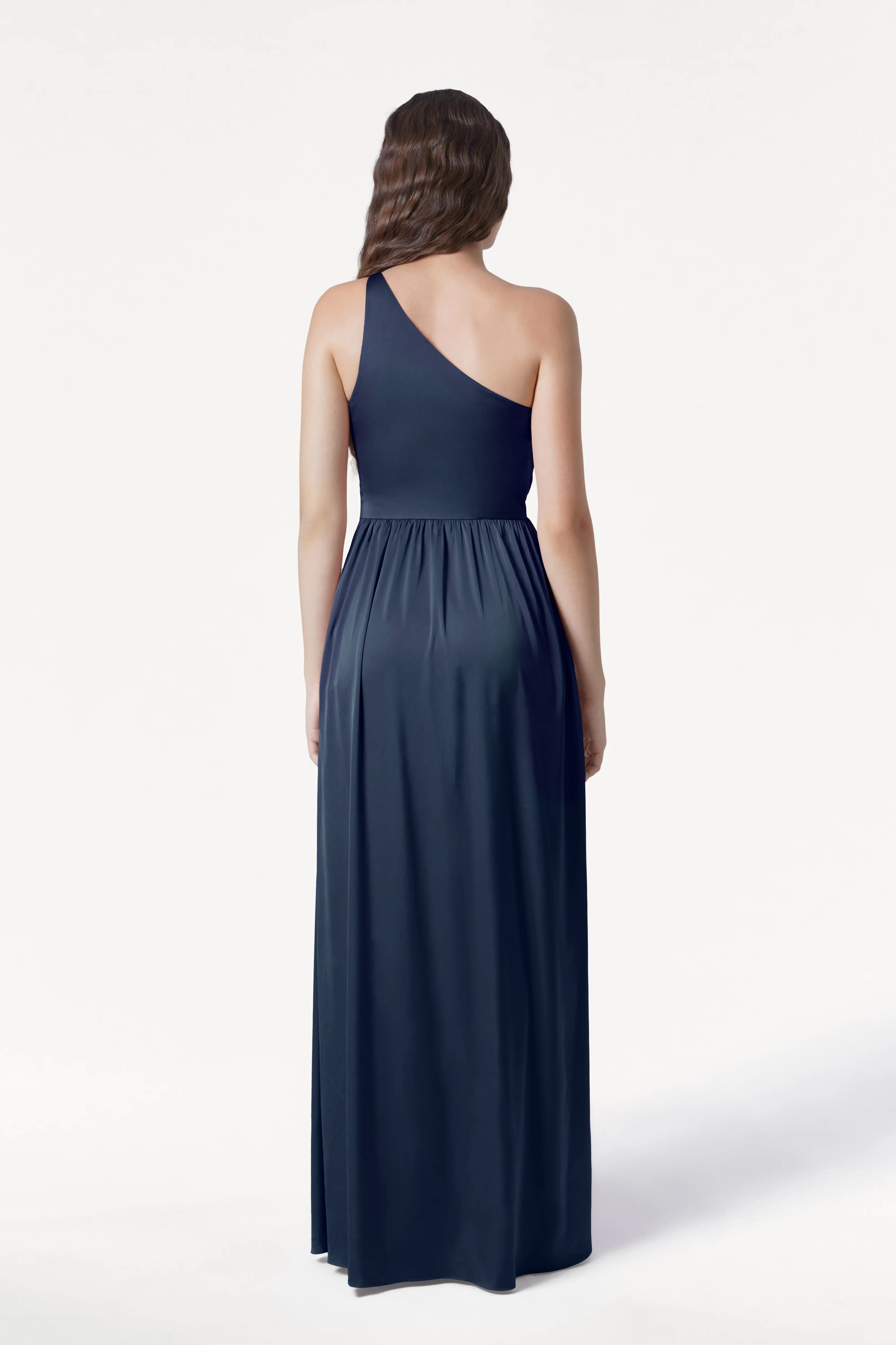 A woman with loose brown waves faces away, modeling the TH&TH Greta Bridesmaid Dress in Navy Satin—a floor-length gown with Grecian draping and a gathered waist—set against a plain white background.