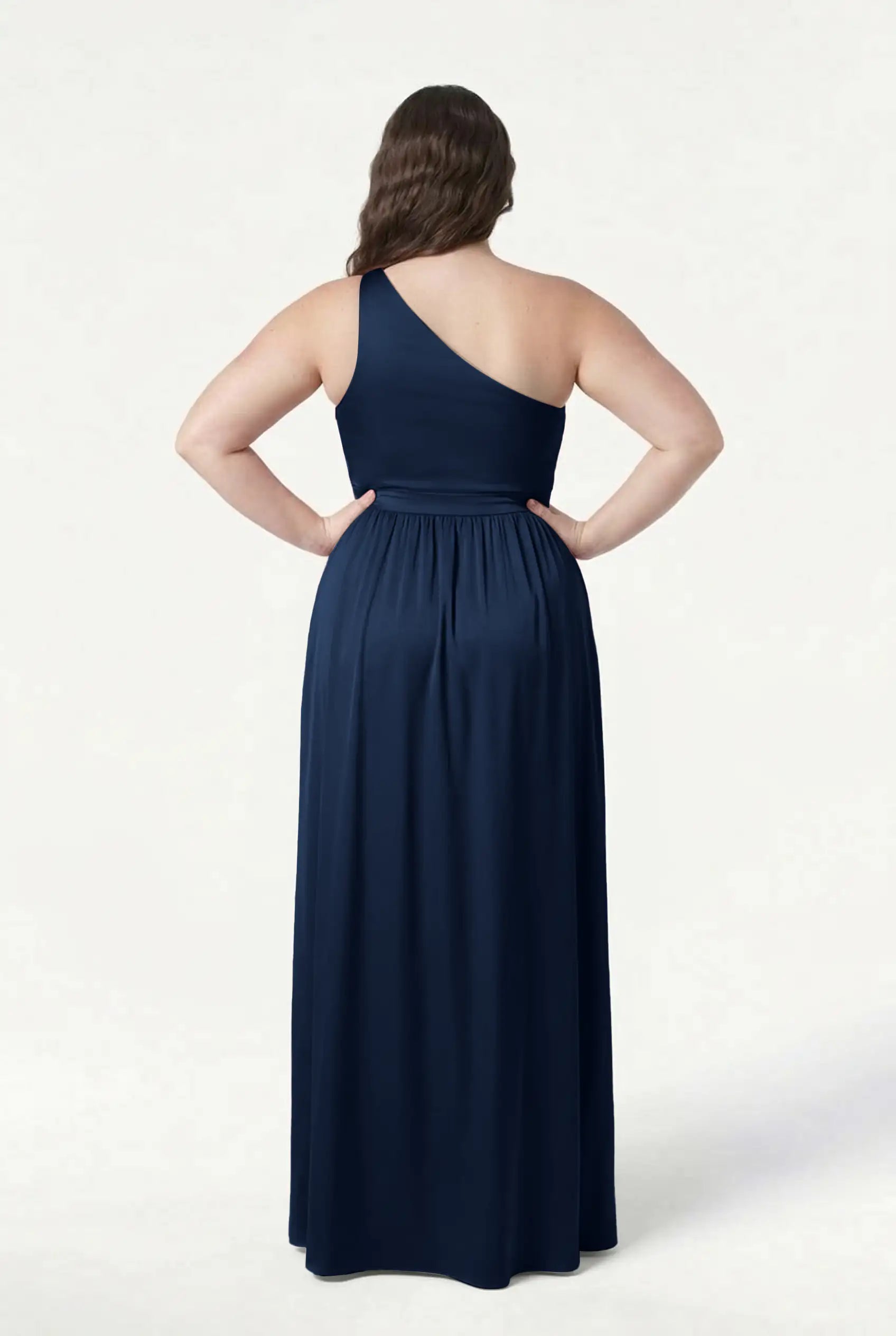A woman stands facing away, hands on hips, in the TH&TH Greta Bridesmaid Dress— a navy satin, one-shoulder, floor-length gown with Grecian draping and a fitted waist, shown against a plain white background.
