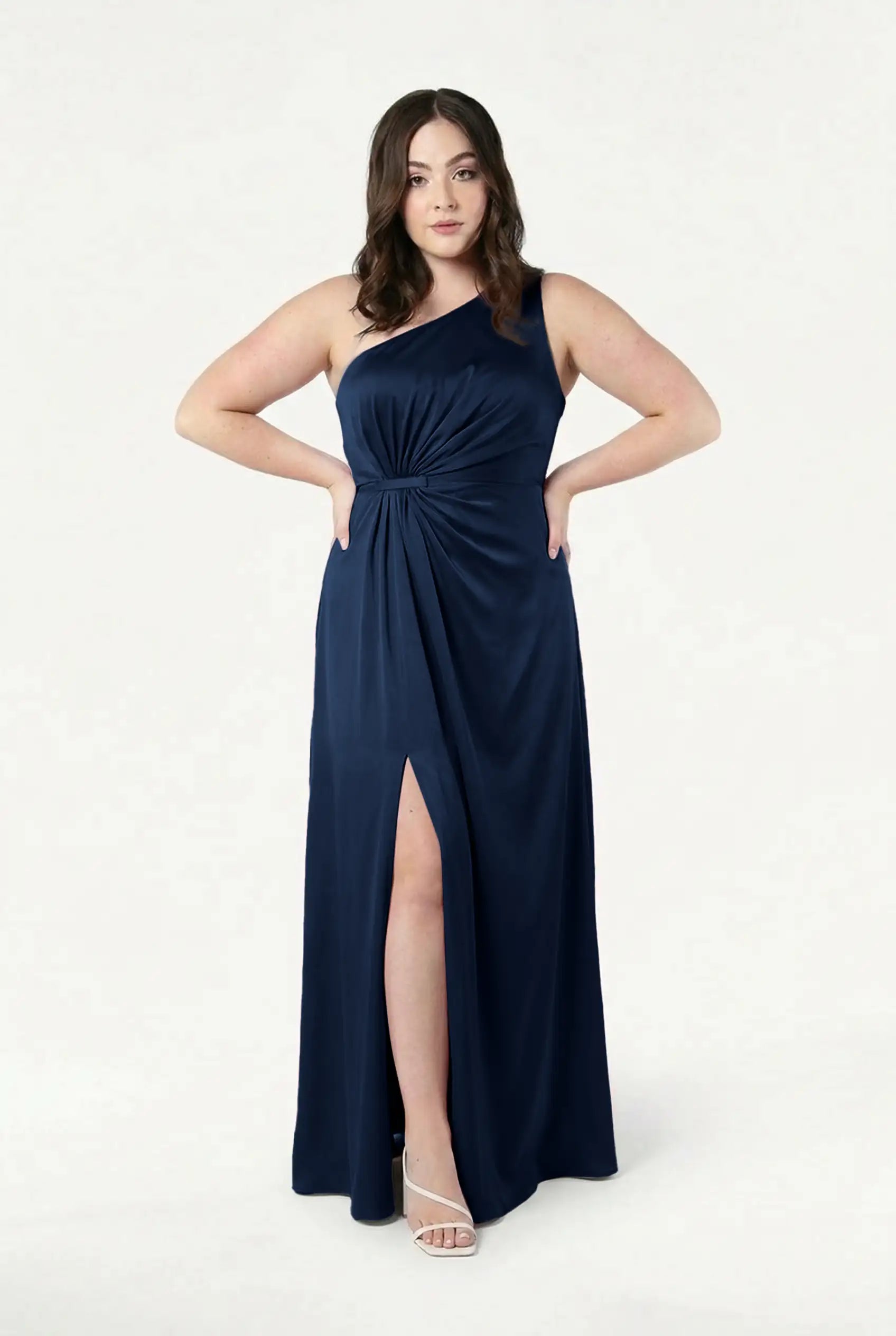 A woman in the TH&TH Greta Bridesmaid Dress in Navy Satin, featuring a one-shoulder Grecian drape, gathered waist knot, and thigh-high slit, stands against a white background with shoulder-length brown hair and light heels.