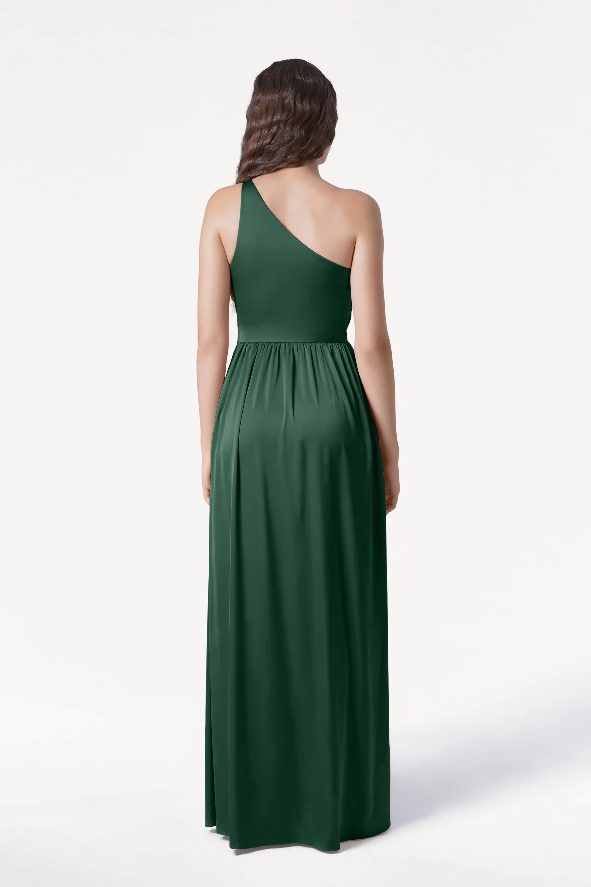 A woman faces away from the camera, showcasing the Greta Bridesmaid Dress in Olive Grove Satin by TH&TH—a floor-length, dark green one-shoulder gown with elegant Grecian draping. Her long, wavy brown hair stands out against a plain white background.