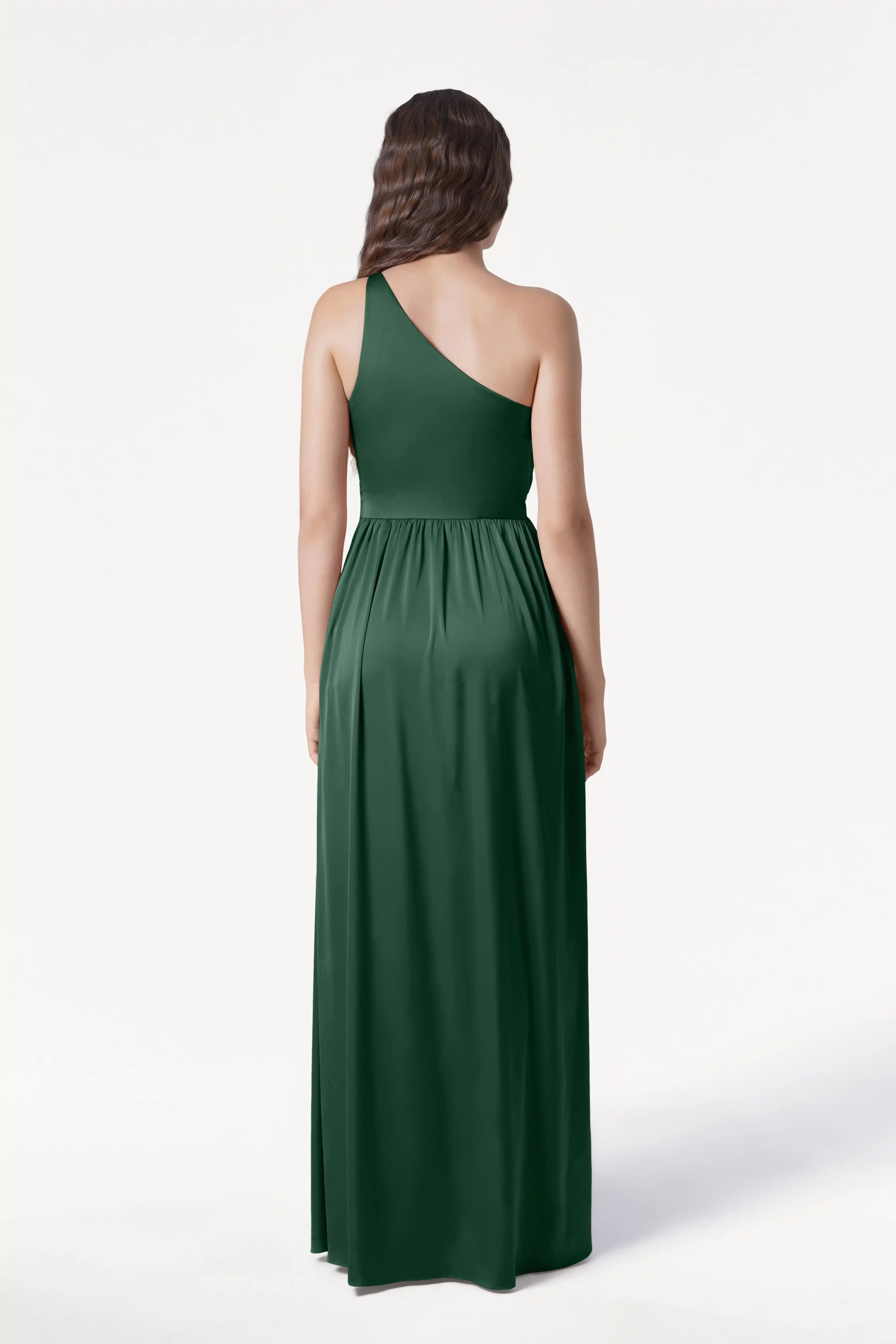 A woman faces away from the camera, showcasing the Greta Bridesmaid Dress in Olive Grove Satin by TH&TH—a floor-length, dark green one-shoulder gown with elegant Grecian draping. Her long, wavy brown hair stands out against a plain white background.