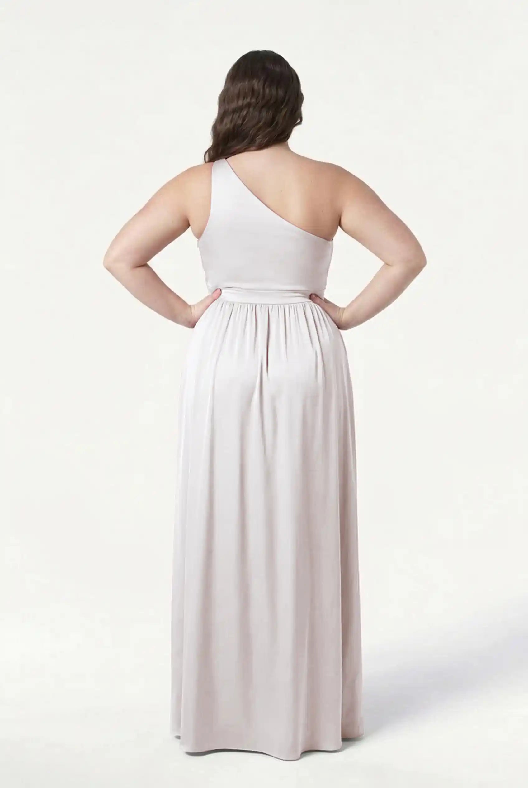 A woman stands with her back to the camera, wearing the TH&TH Greta Bridesmaid Dress in Oyster Satin. This long, one-shoulder gown features Grecian draping, a fitted waist, and a flowing floor-length skirt against a plain white background.